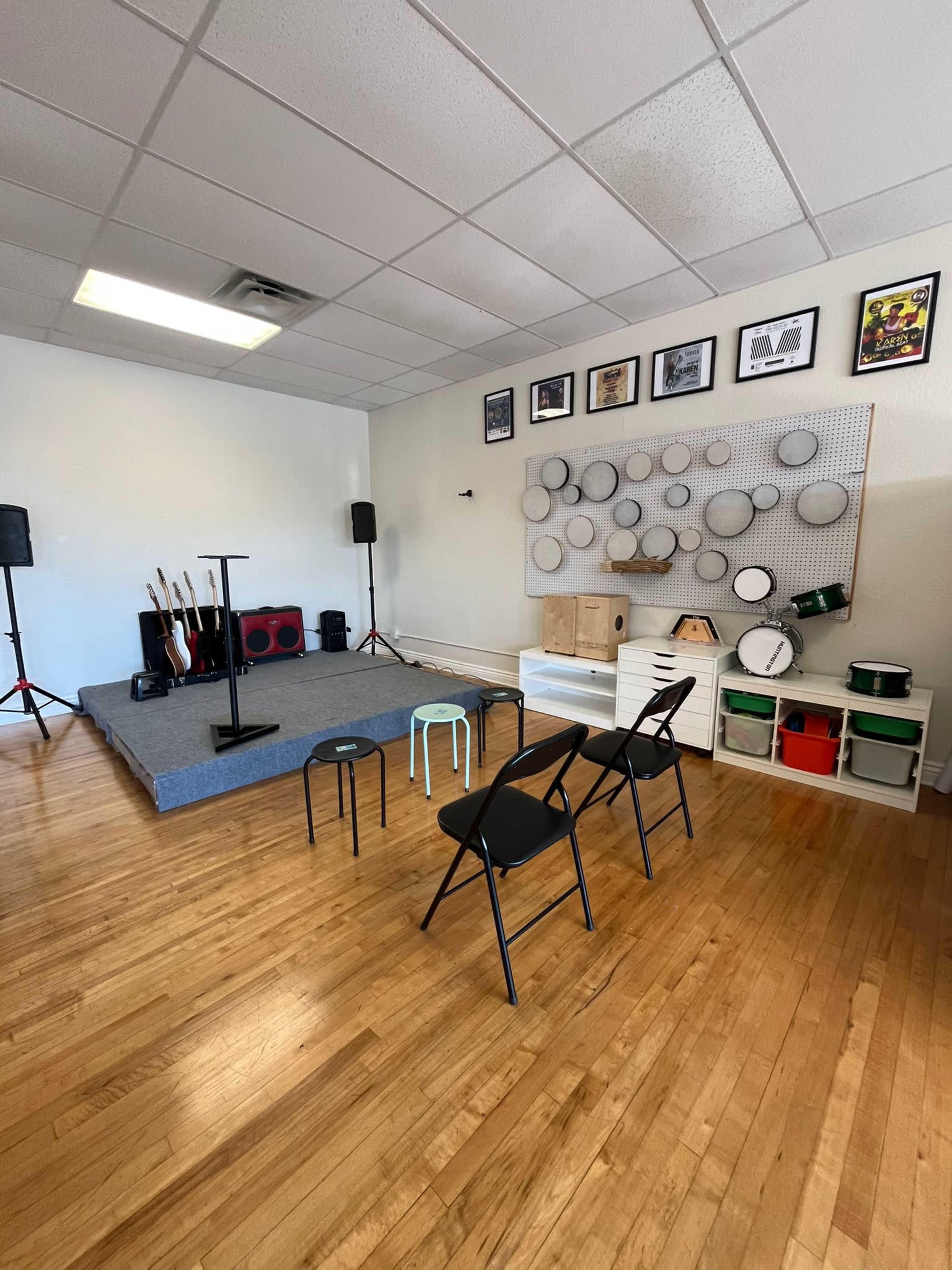 The image shows a music practice room with a small stage, several musical instruments, a pegboard with drum circles, and chairs arranged in front of the stage.