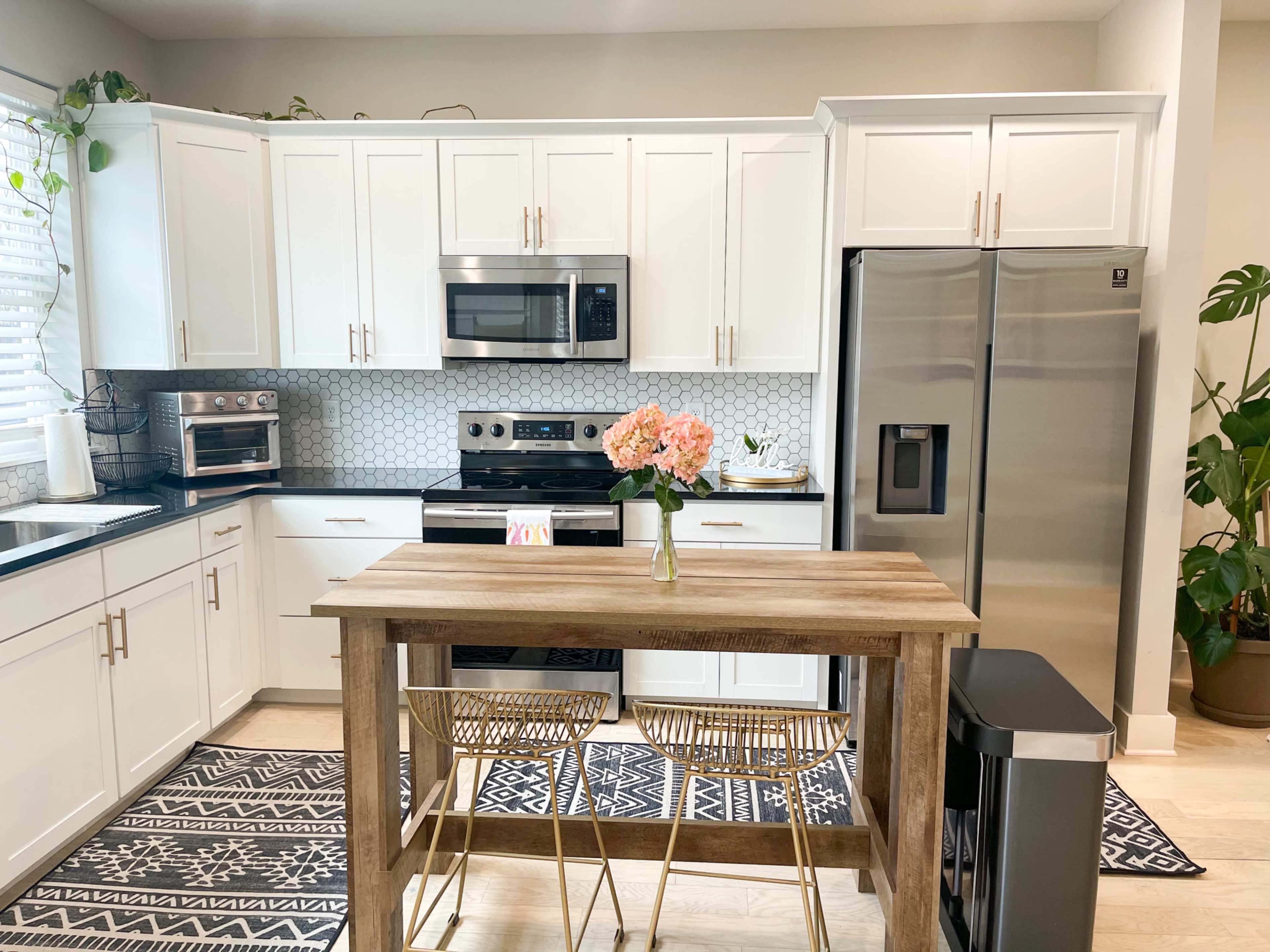 A modern kitchen features white cabinets, stainless steel appliances, and a wooden table with two chairs, complemented by a decorative plant and flowers.