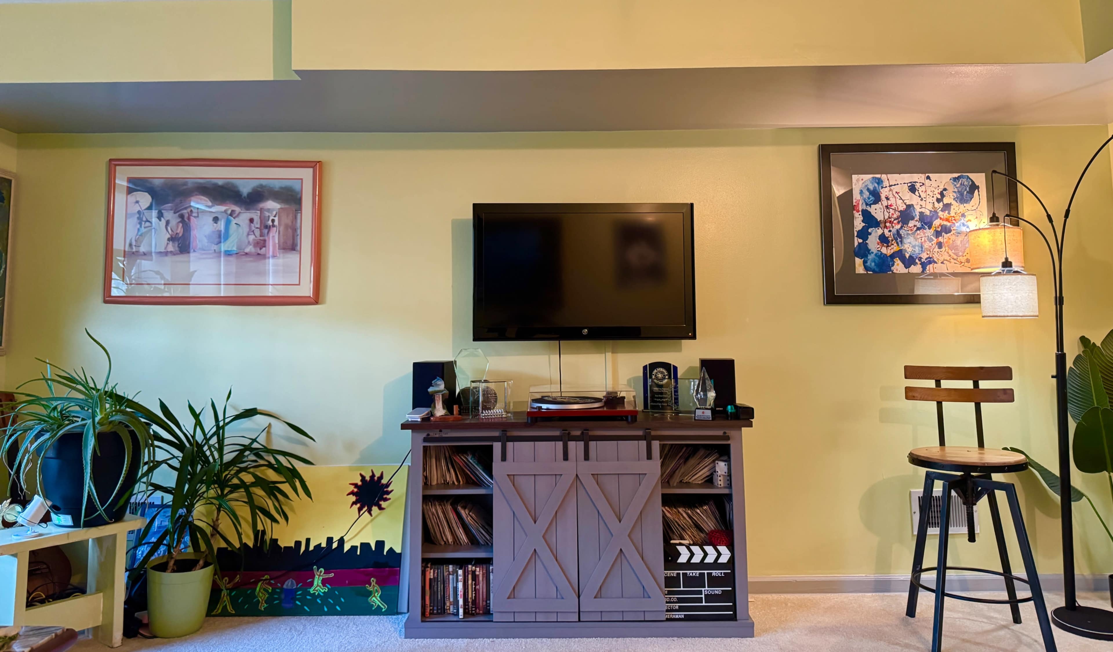 The image shows a living room with a television mounted on a yellow wall, flanked by framed artwork, a wooden entertainment center, and a high stool beside a potted plant.