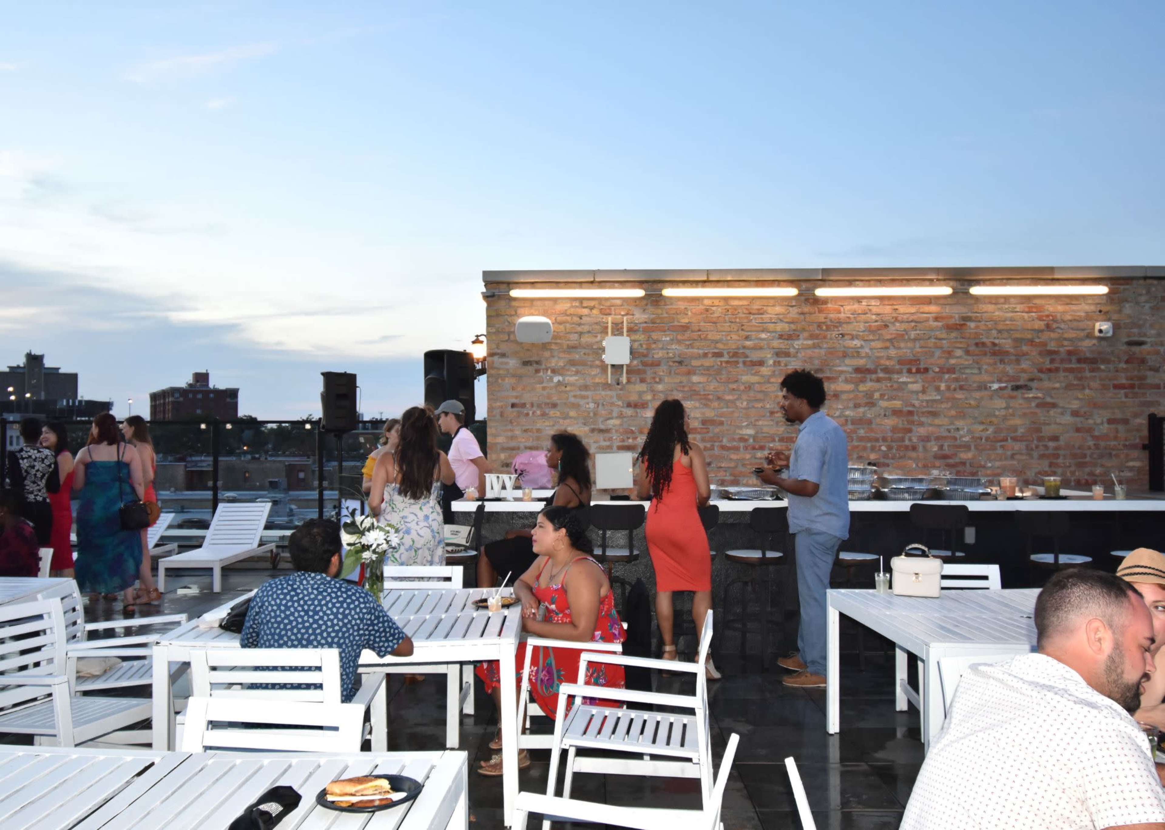 People socialize on a rooftop with white furniture and a city skyline in the background during an evening event.