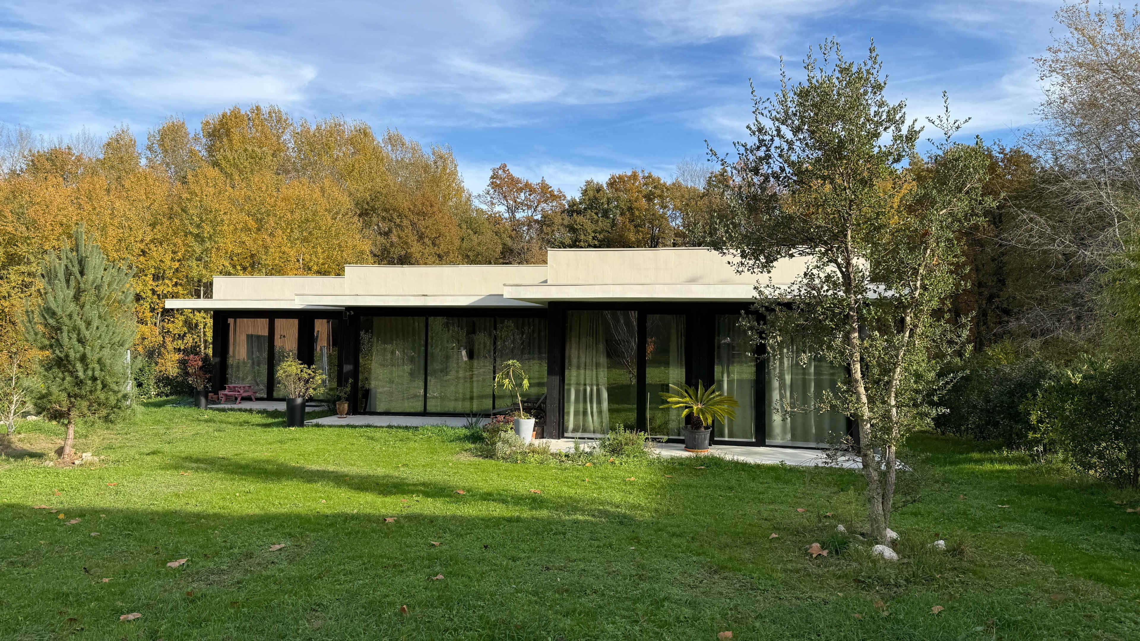 A modern single-story house with large glass windows is surrounded by greenery and autumn-colored trees.