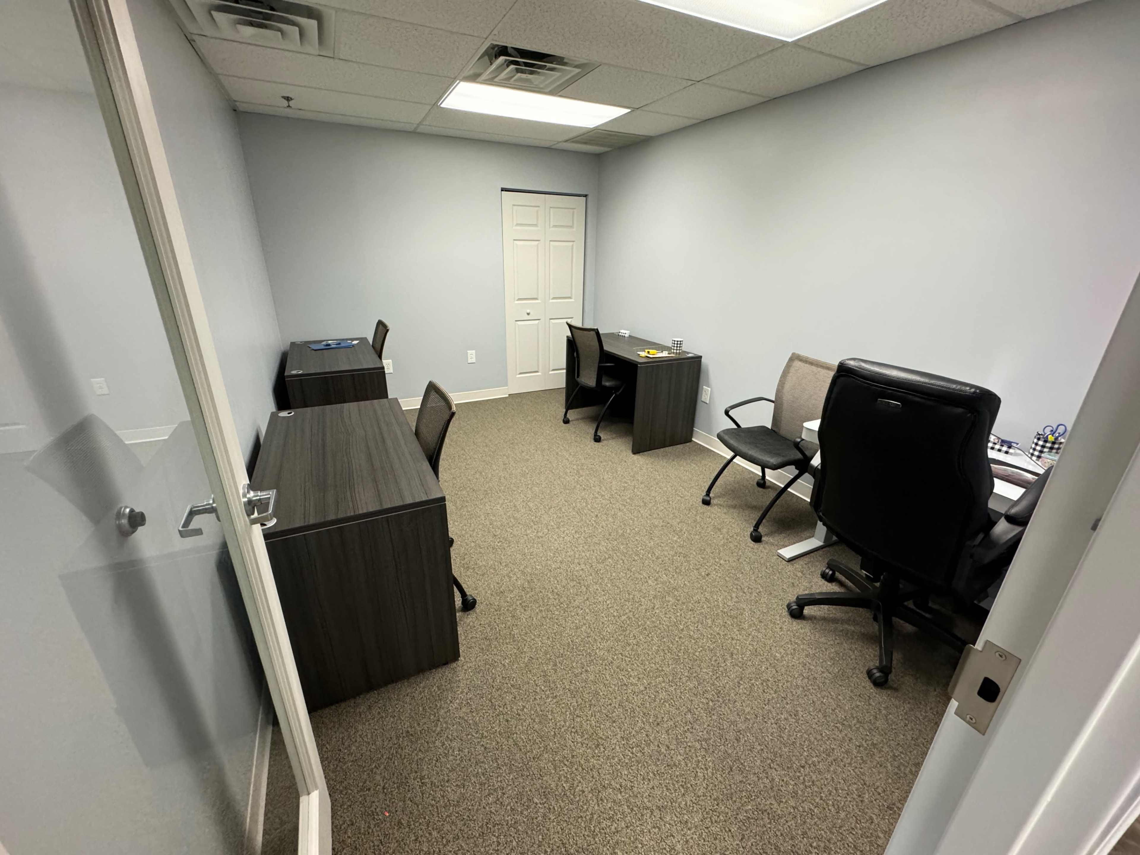 A vacant office space with two desks and chairs on either side, and a closed door at the back.