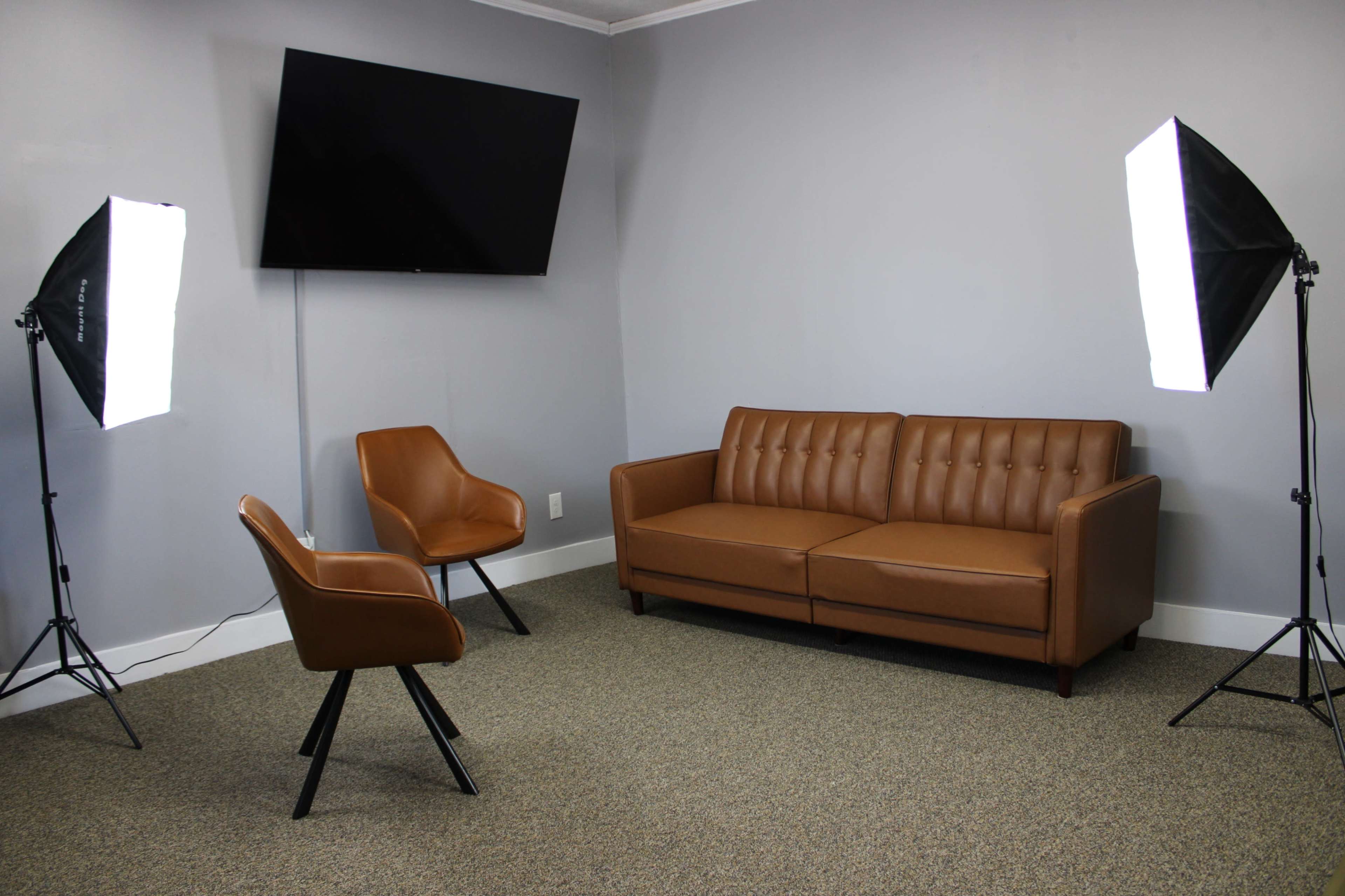 The image shows a small, minimalist room featuring a brown leather sofa, a matching chair, and two lighting stands, with a wall-mounted television in the background.