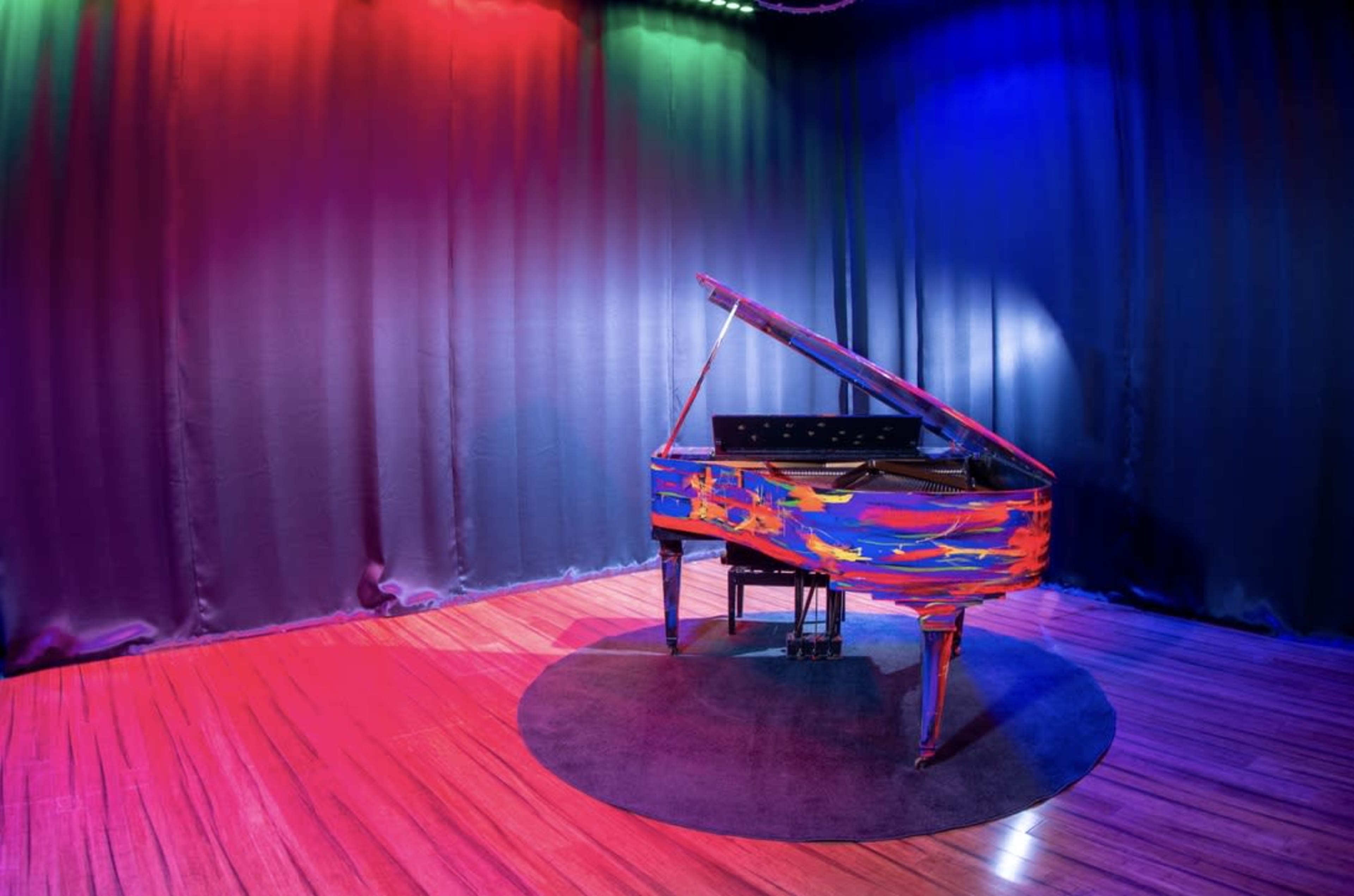 A brightly painted grand piano sits on a circular rug in a stage area illuminated by colorful lights against a dark backdrop.