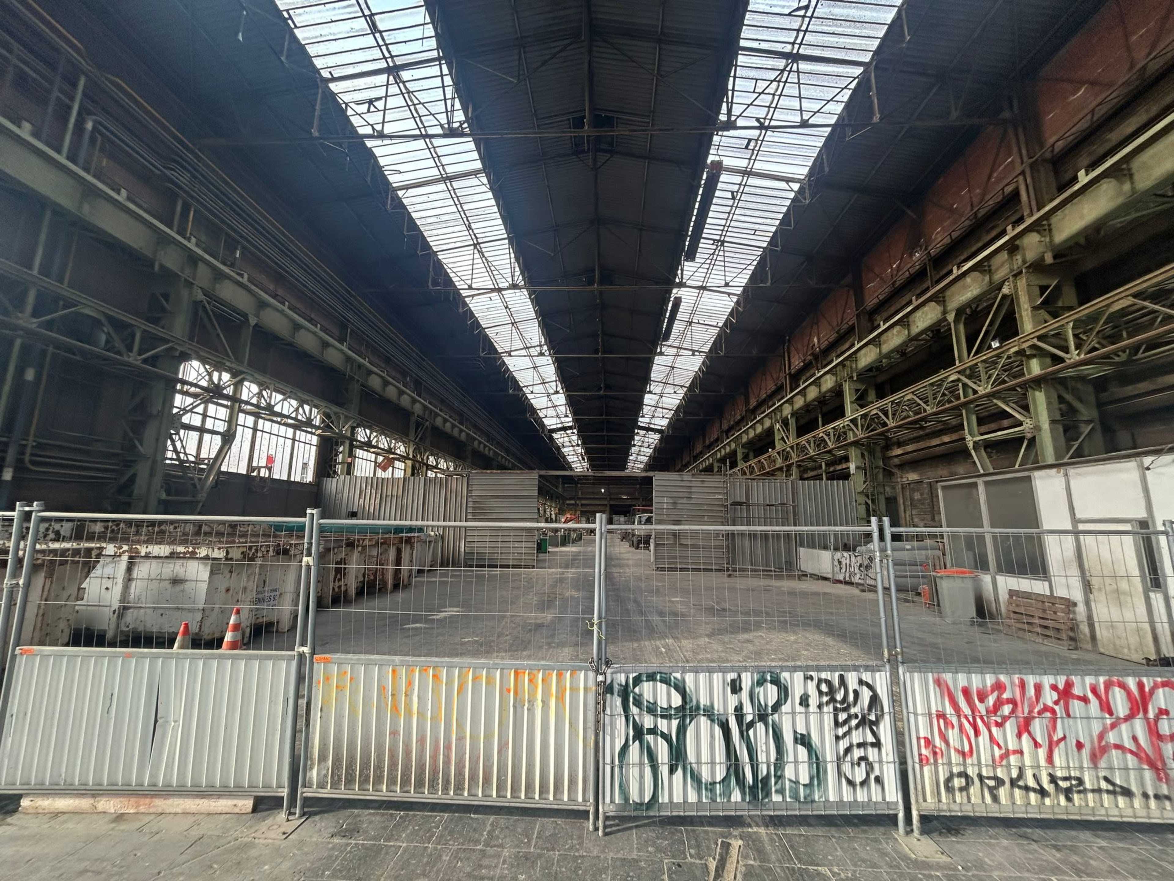 An industrial building interior features a large open space, surrounded by metal fencing, with graffiti on the walls and skylights above.