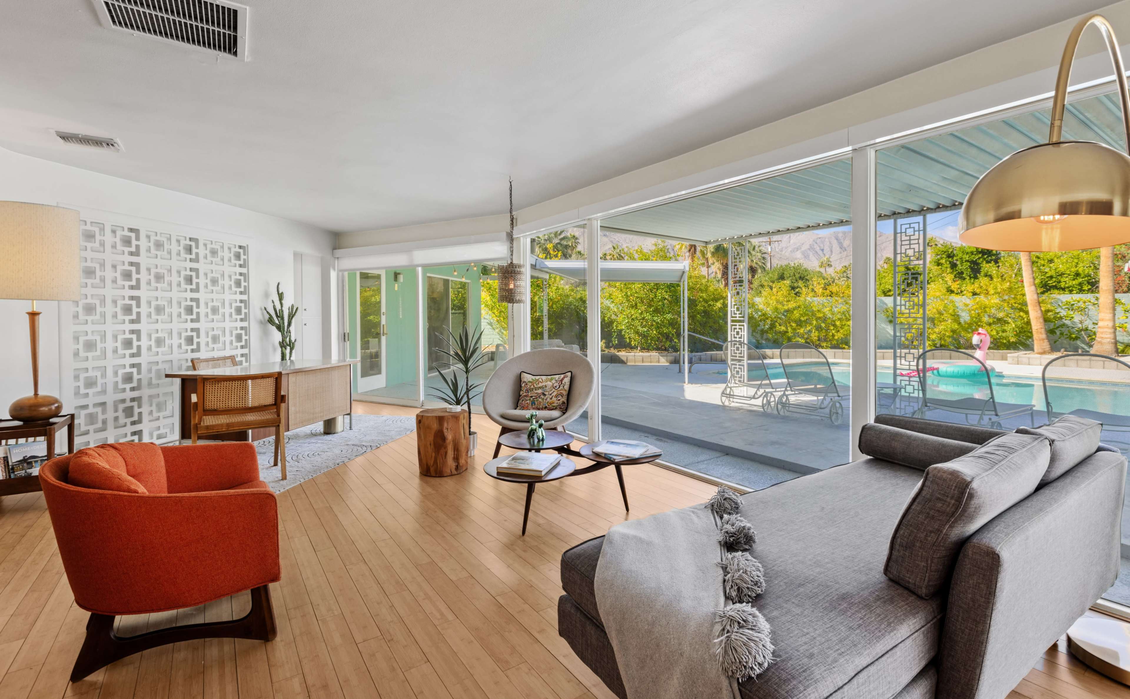 The image shows a modern living room with large glass windows overlooking a patio and pool area, featuring minimalist furniture and a light color palette.