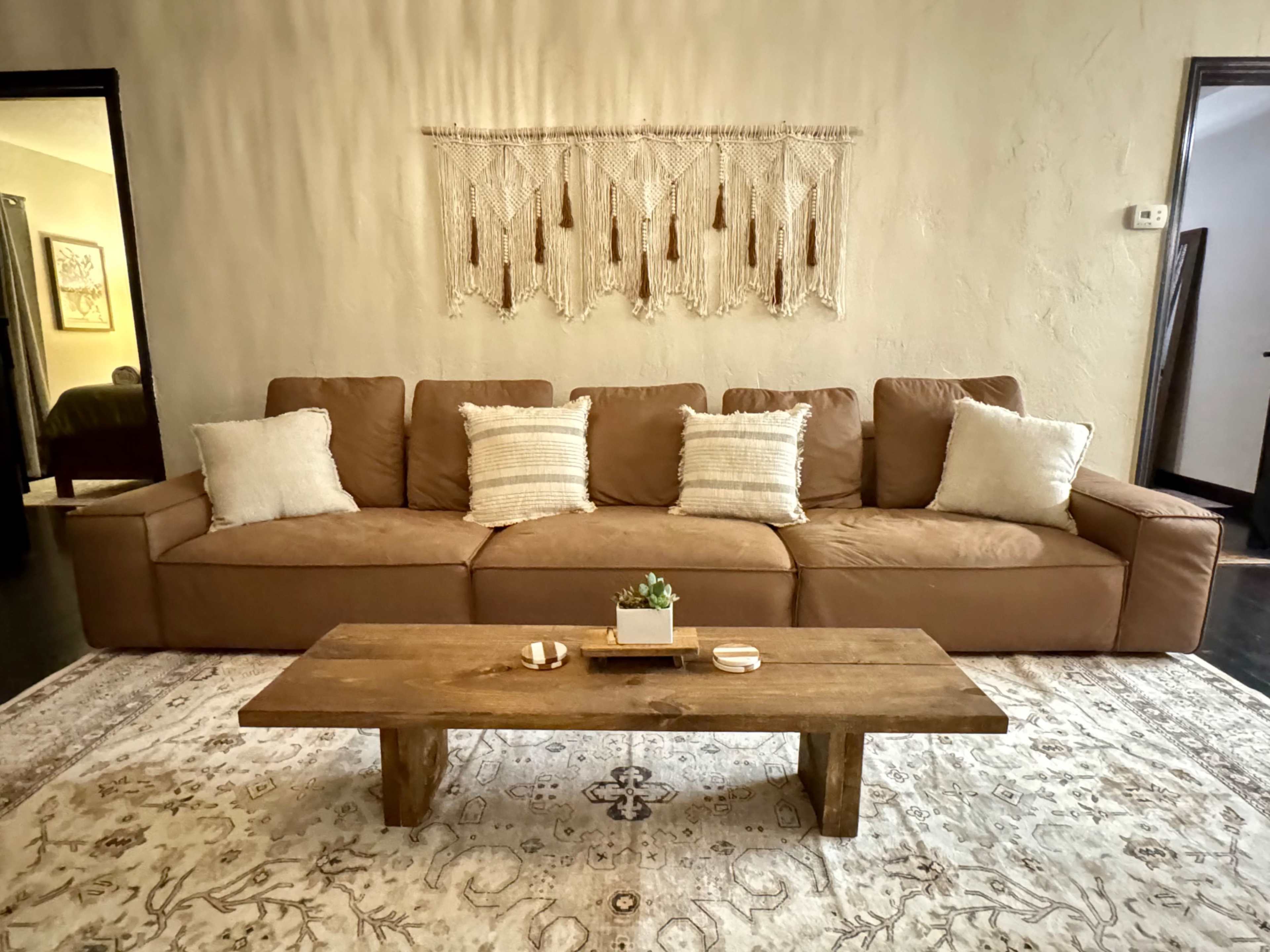 The image shows a cozy living room with a brown sectional sofa, a rustic wooden coffee table, and a decorative wall hanging above the sofa.