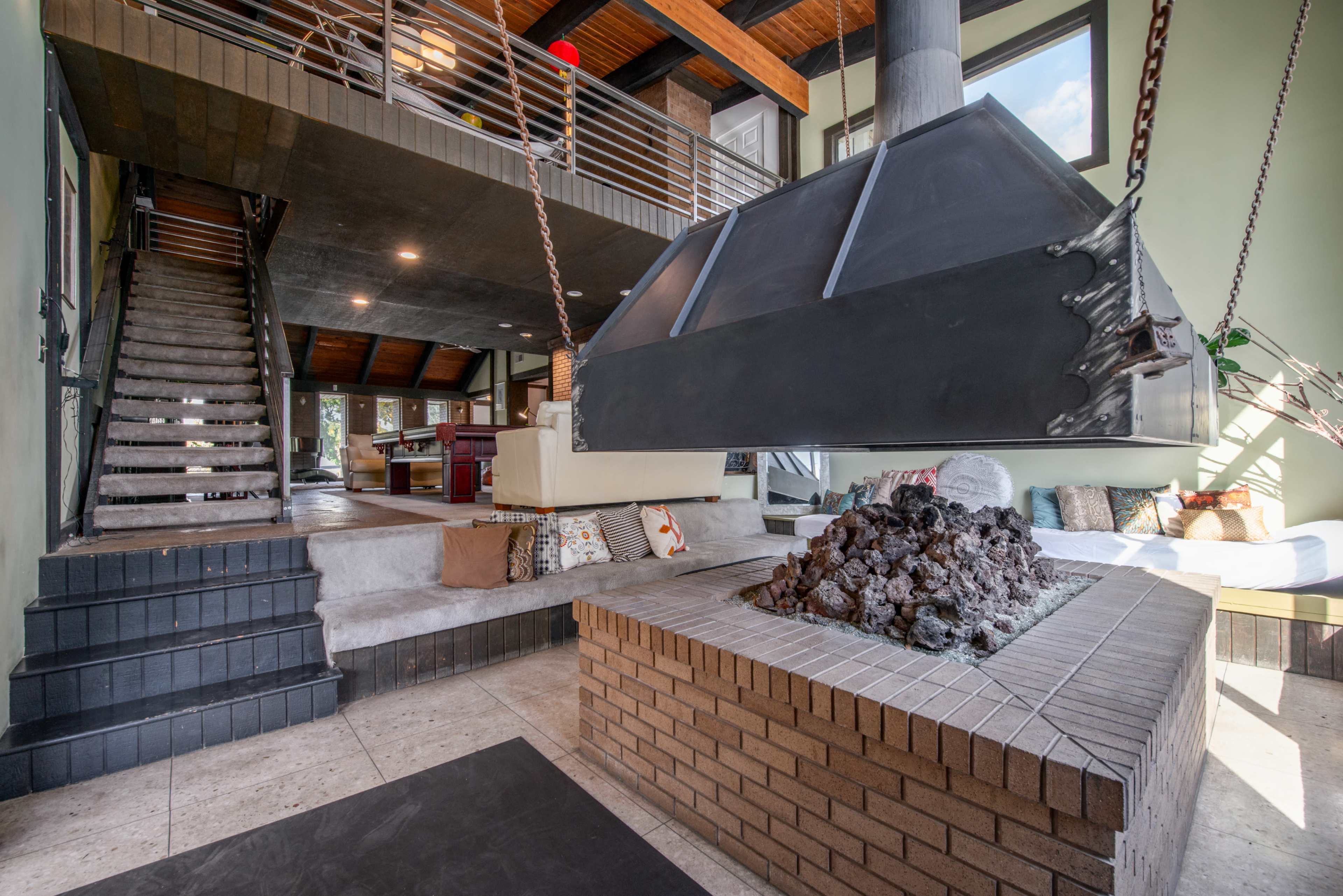 A modern living space with a suspended fireplace at the center, surrounded by a brick seating area and a staircase leading to an upper level.