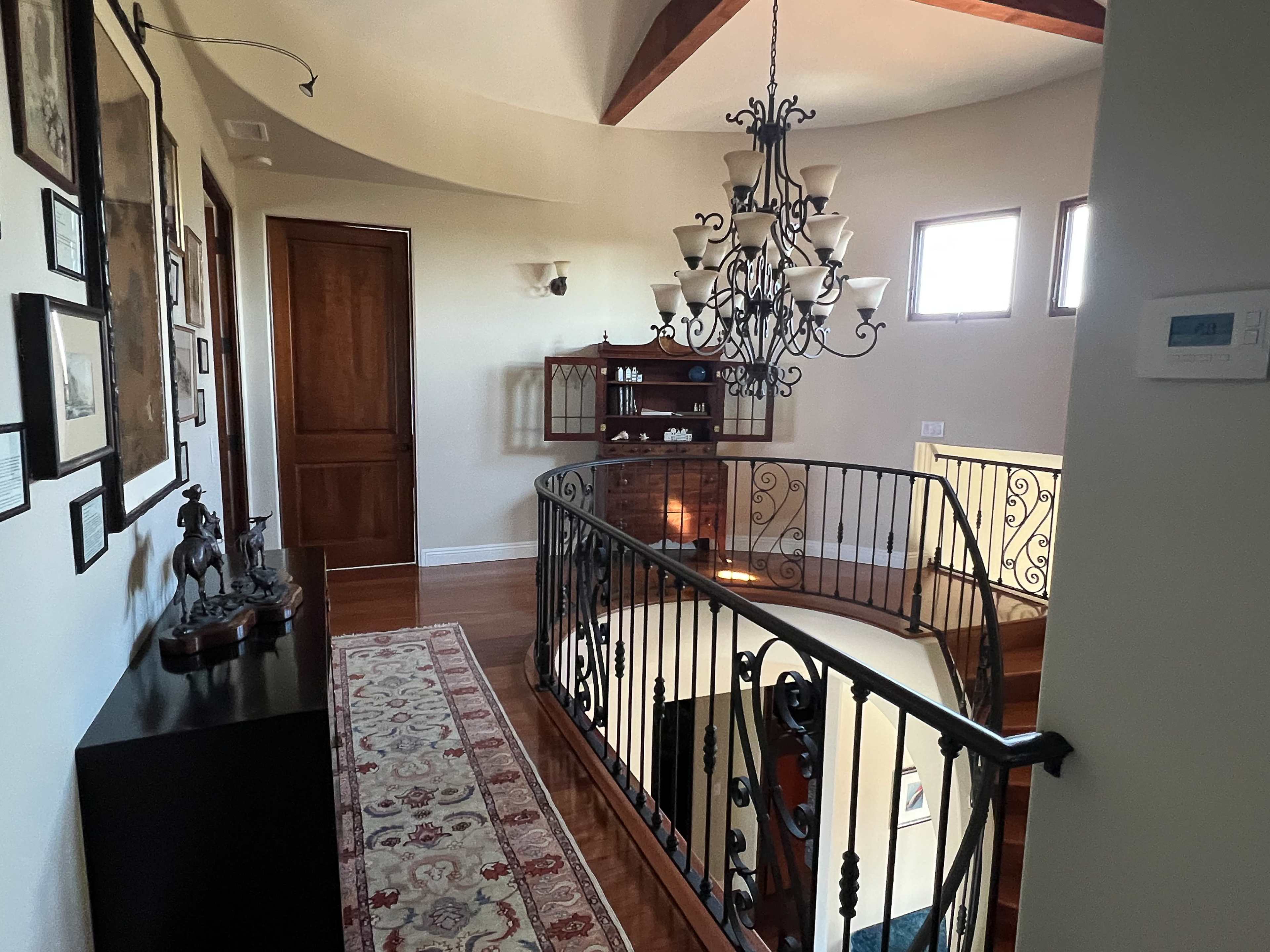 The image shows a hallway with a curved staircase, a wooden door, a chandelier, wall art, and a small shelf against the wall.
