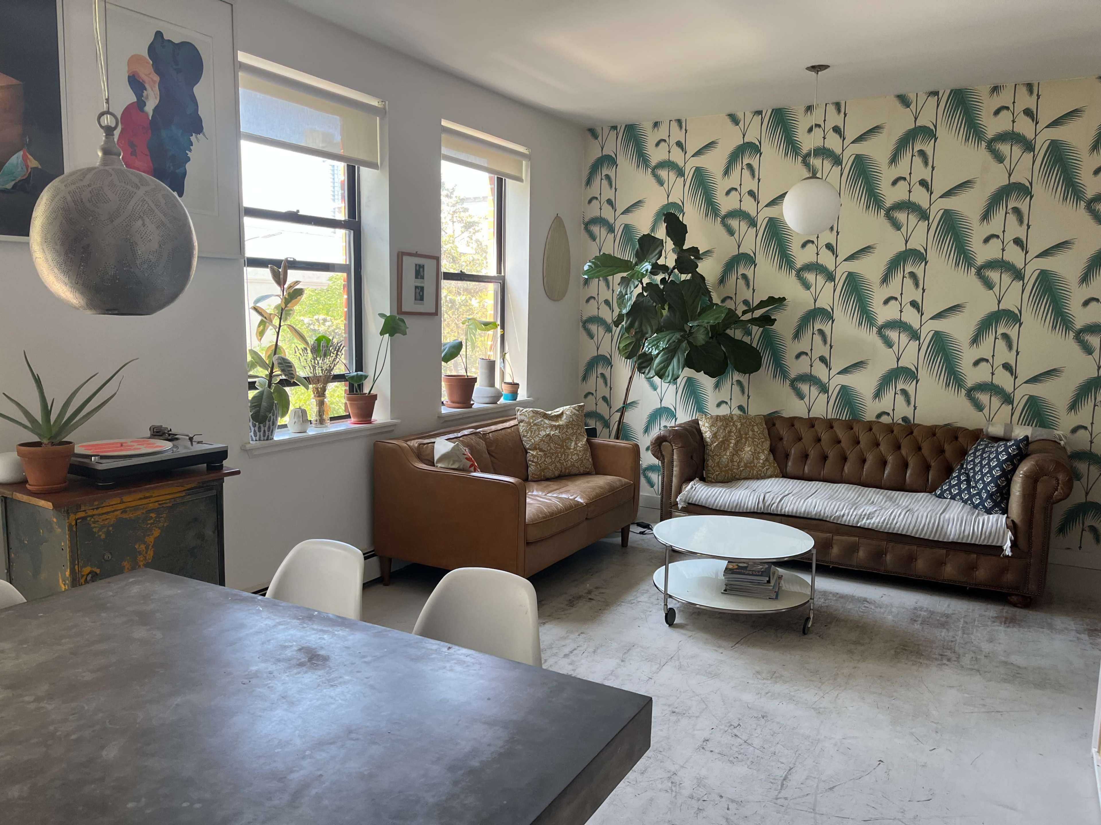 The image shows a living room featuring a brown leather couch, a white coffee table, large windows with plants, and a wall decorated with tropical-themed wallpaper.