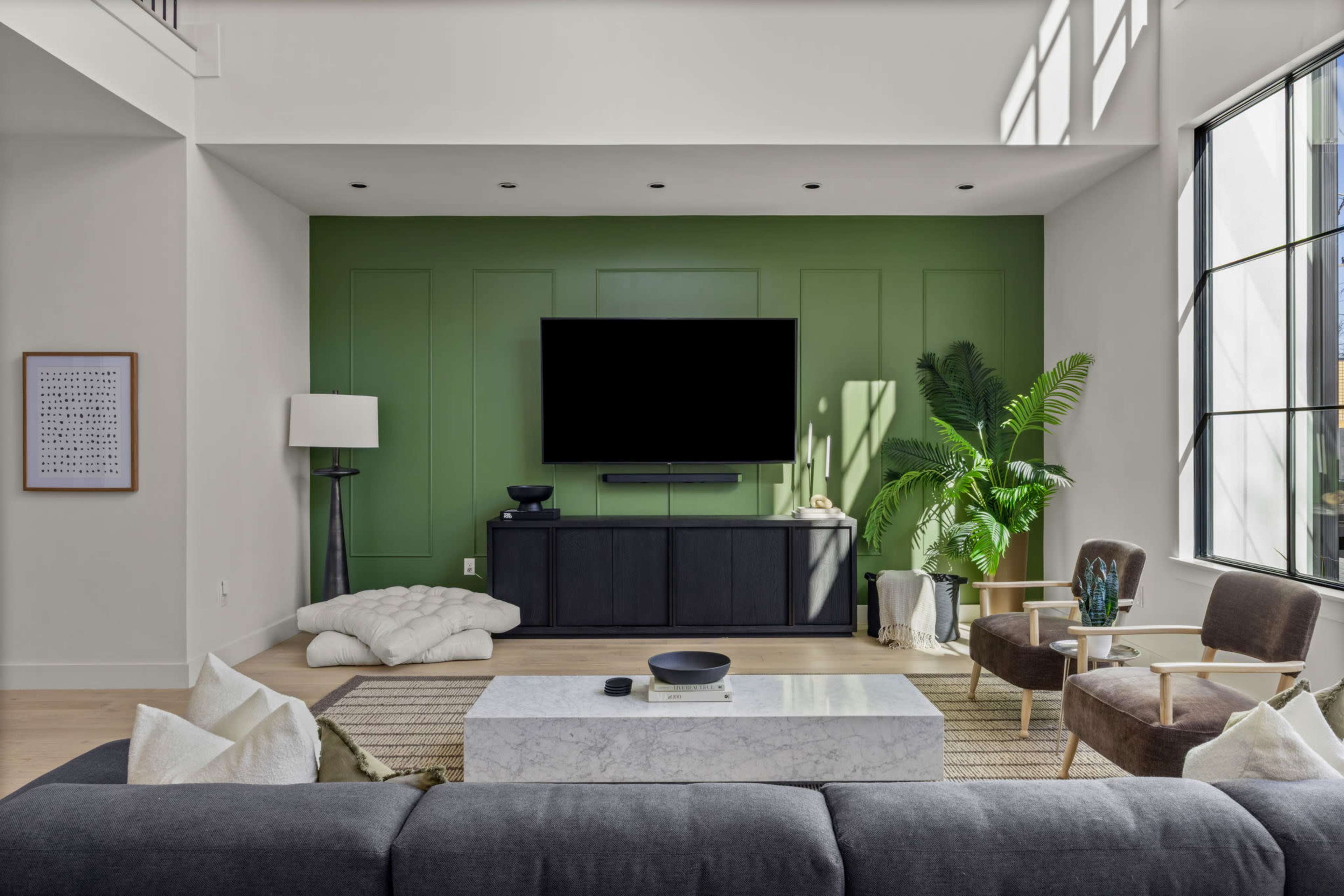 A modern living room features a green accent wall with a mounted television, a marble coffee table, and plants, complemented by cozy seating arrangements.