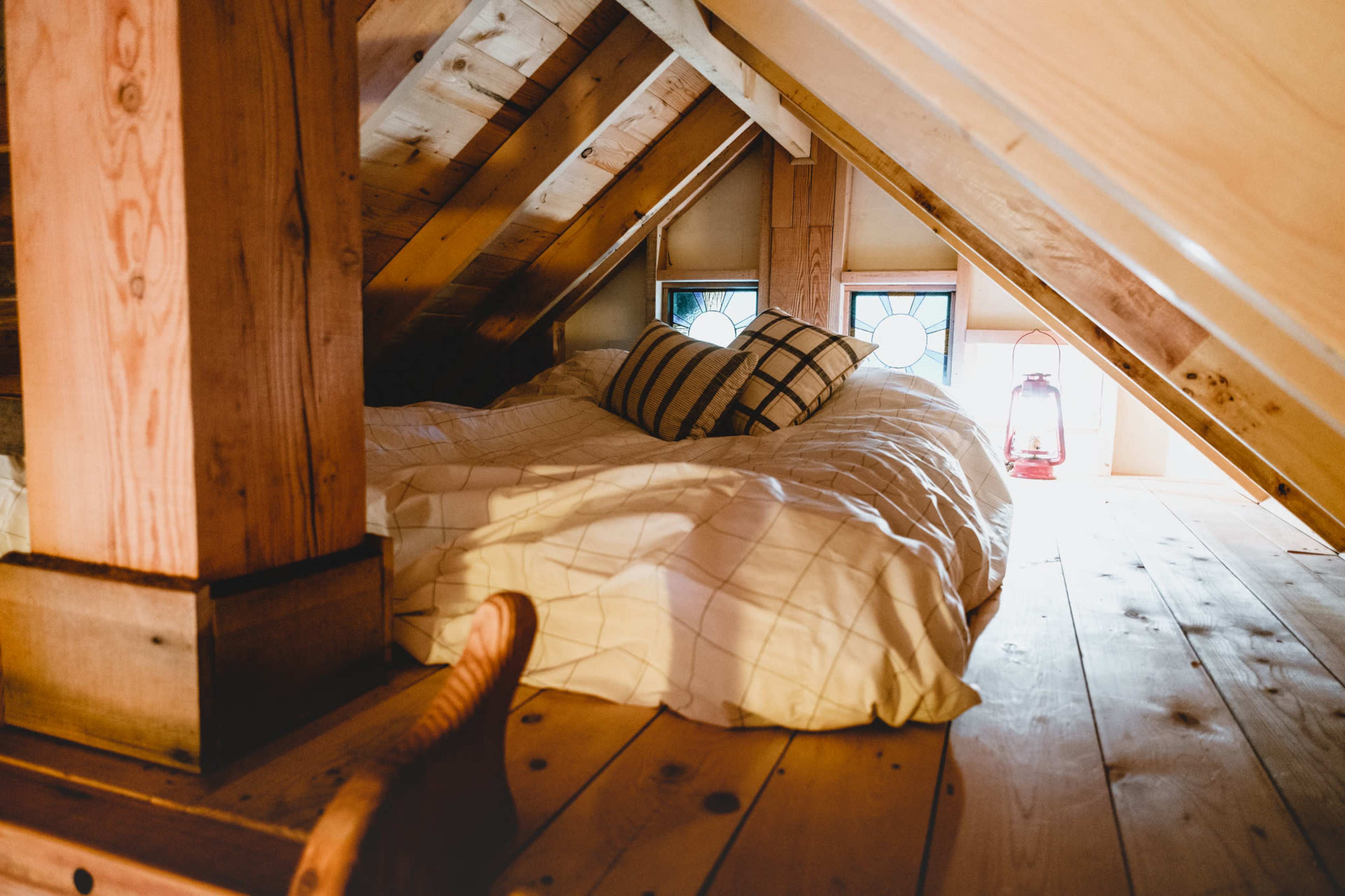 A cozy attic space features a bed with white bedding and decorative pillows, surrounded by wooden beams and a lantern by the window.