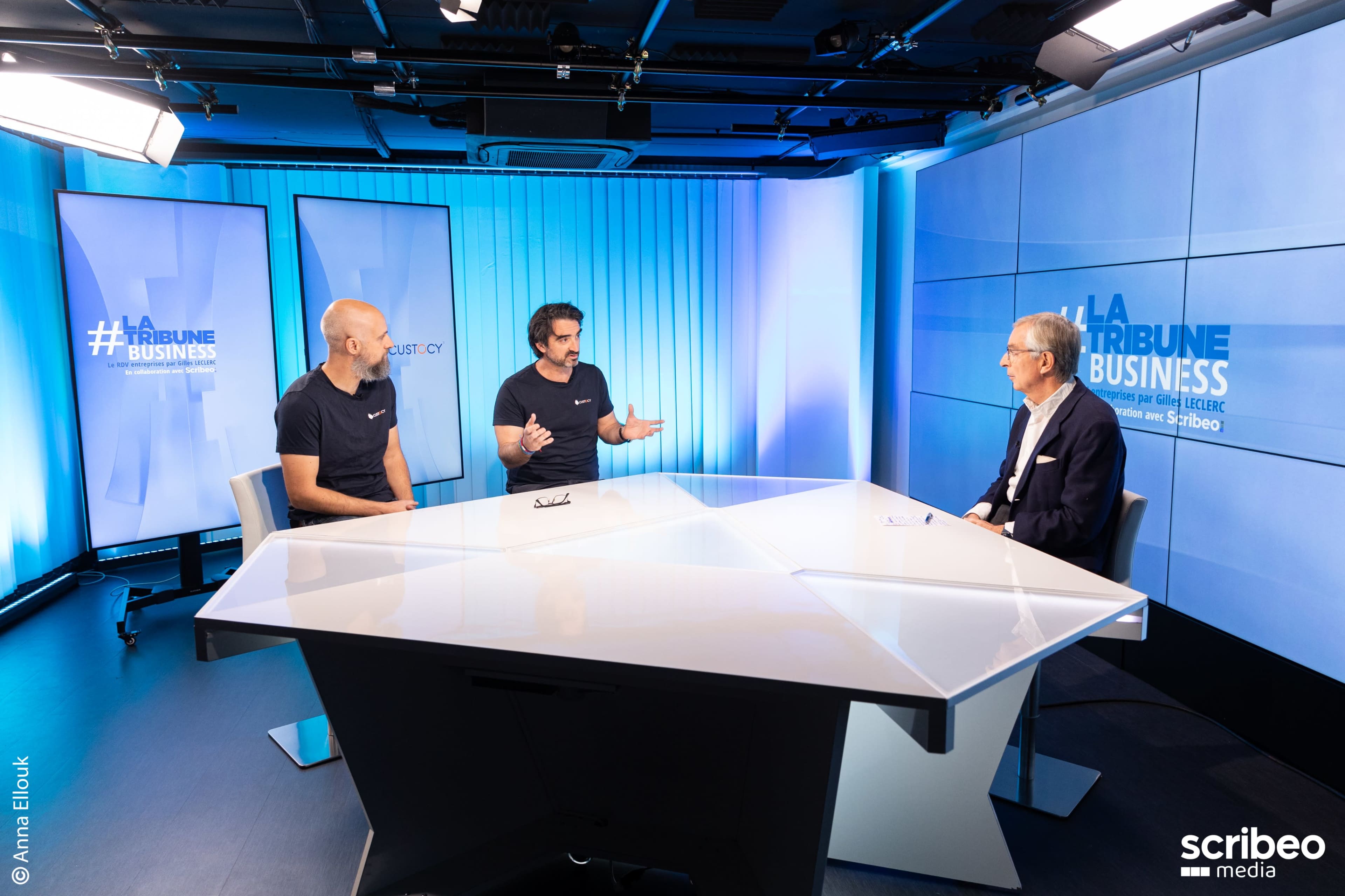 Three men are seated at a white table in a studio, discussing in front of screens displaying "LA TRIBUNE BUSINESS."
