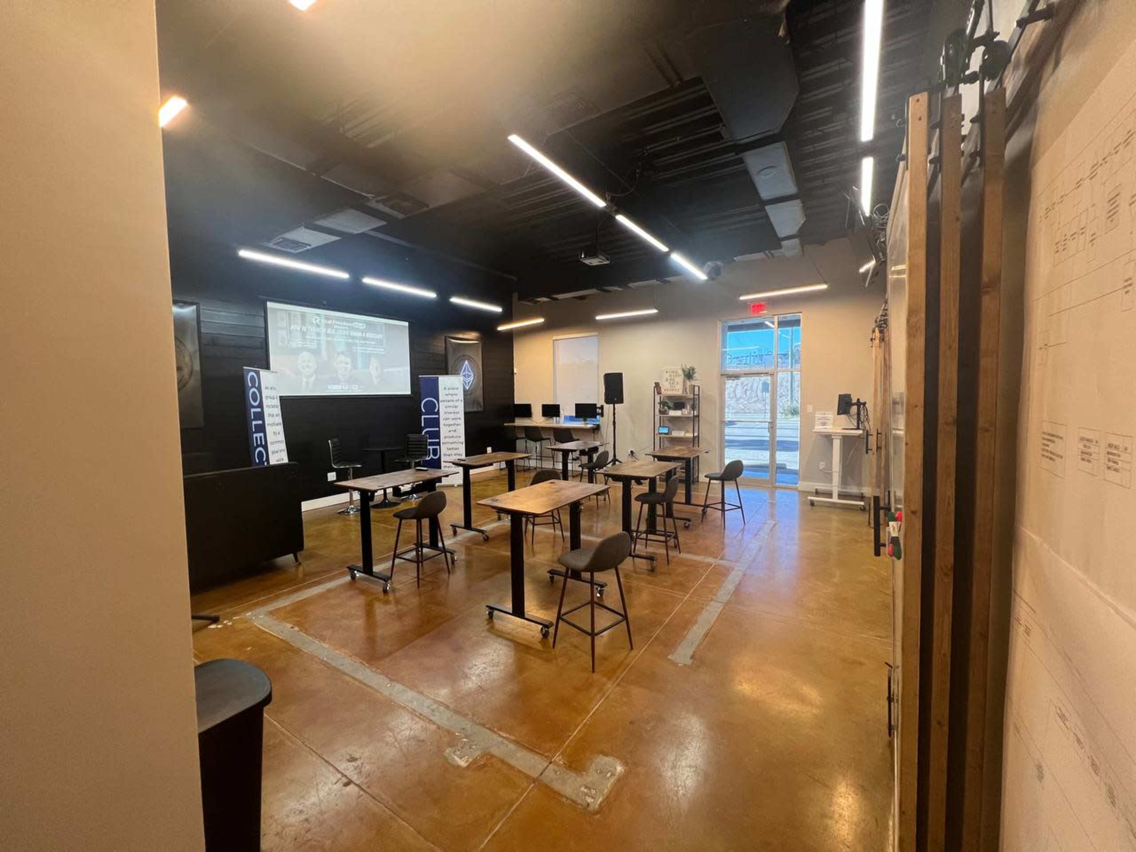 A modern classroom or meeting space with tables, chairs, and a presentation screen along one wall.