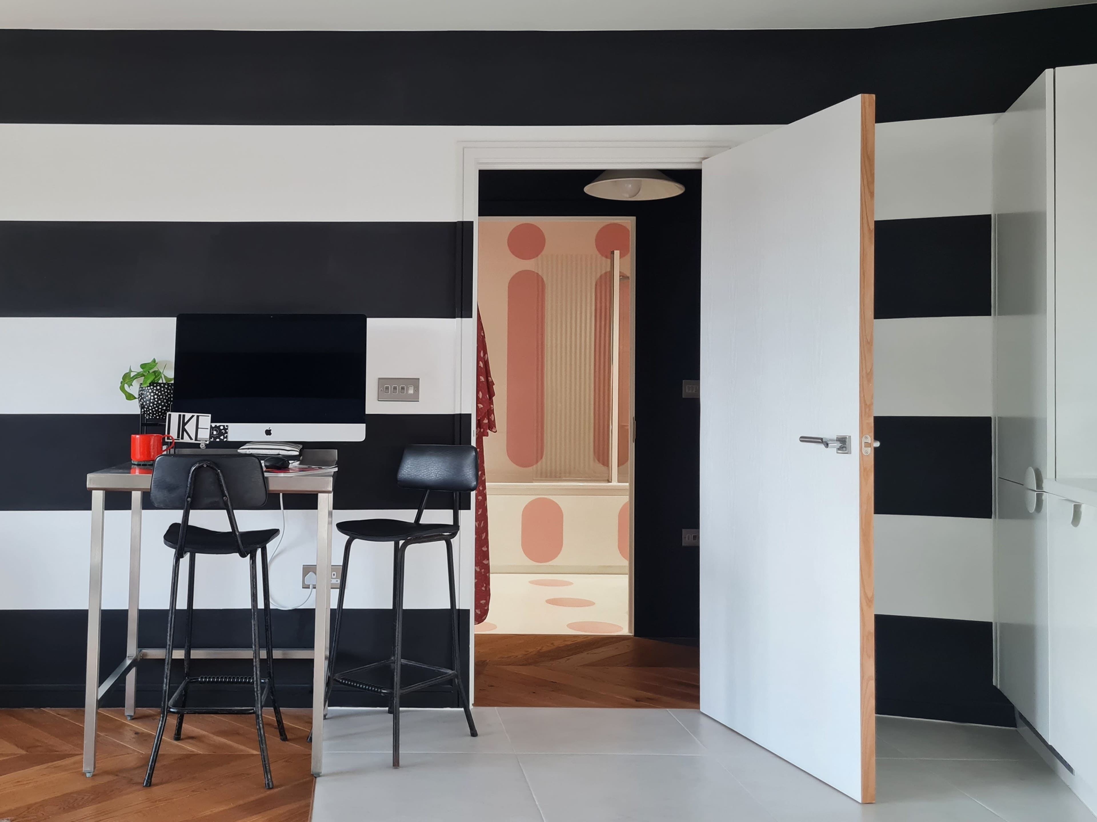A modern kitchen features a black and white striped wall, a small table with two stools, and an open door leading to a room with a patterned floor.