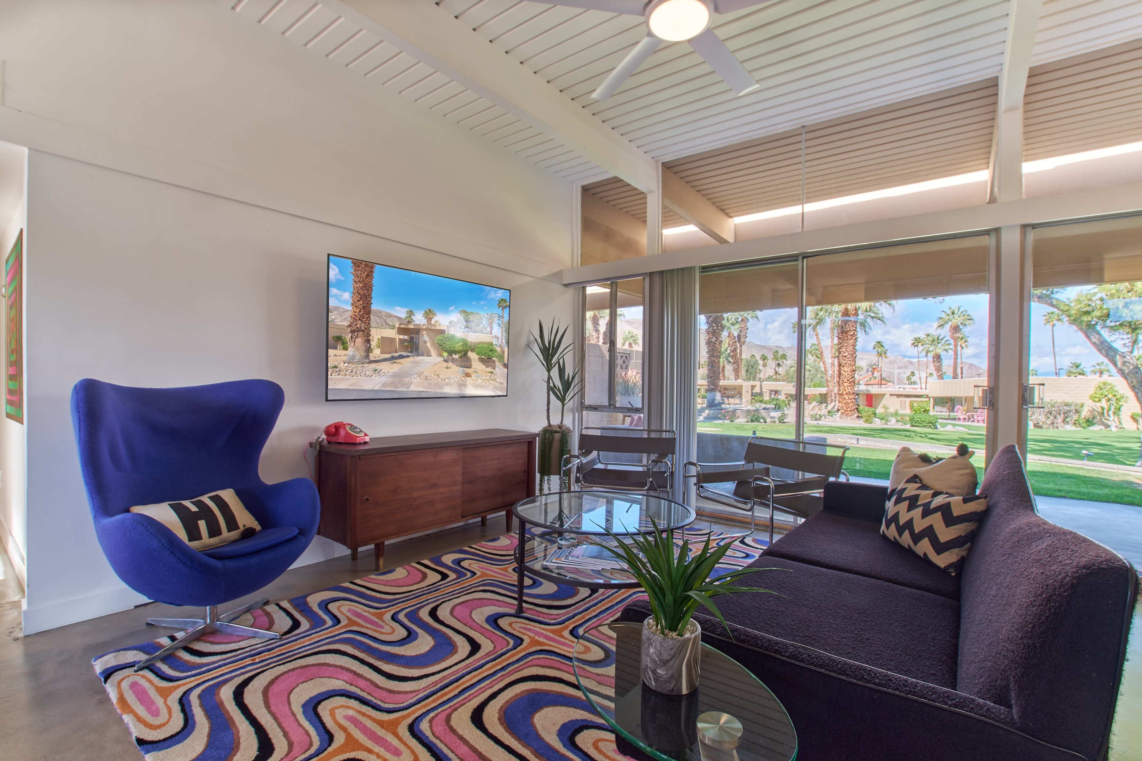 A modern living room features a blue accent chair, a glass coffee table, and a large window with a view of palm trees and outdoor space.