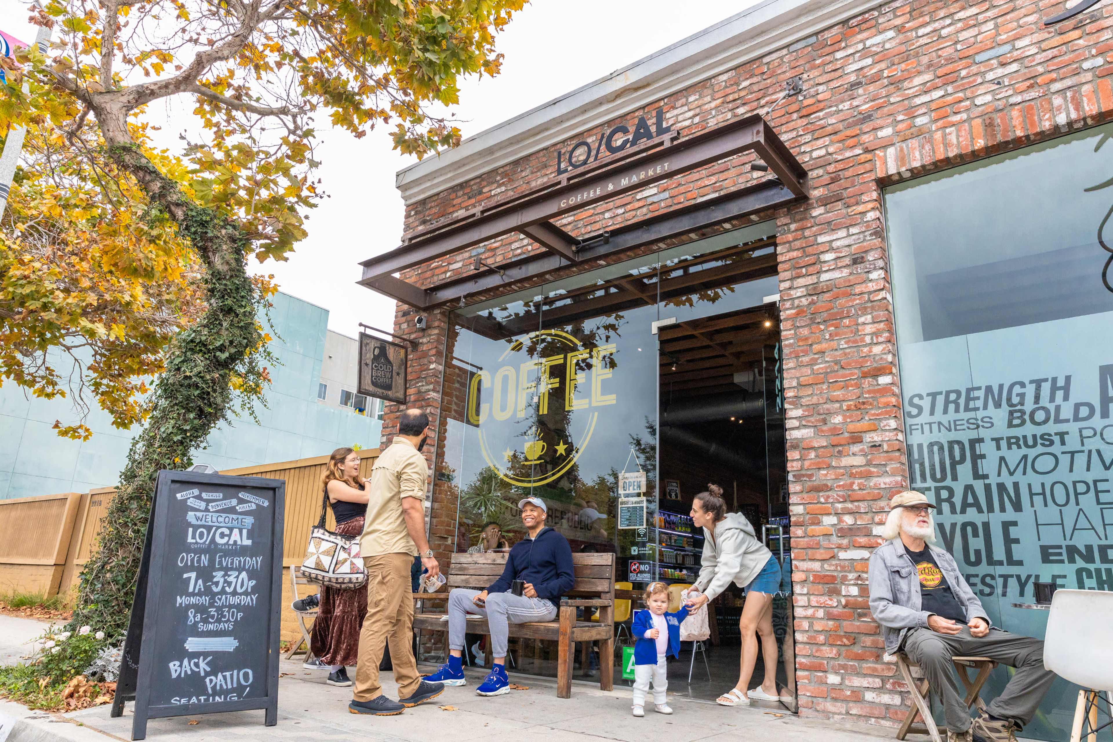 A small coffee shop with a brick facade features people sitting outside and a sign indicating opening hours.