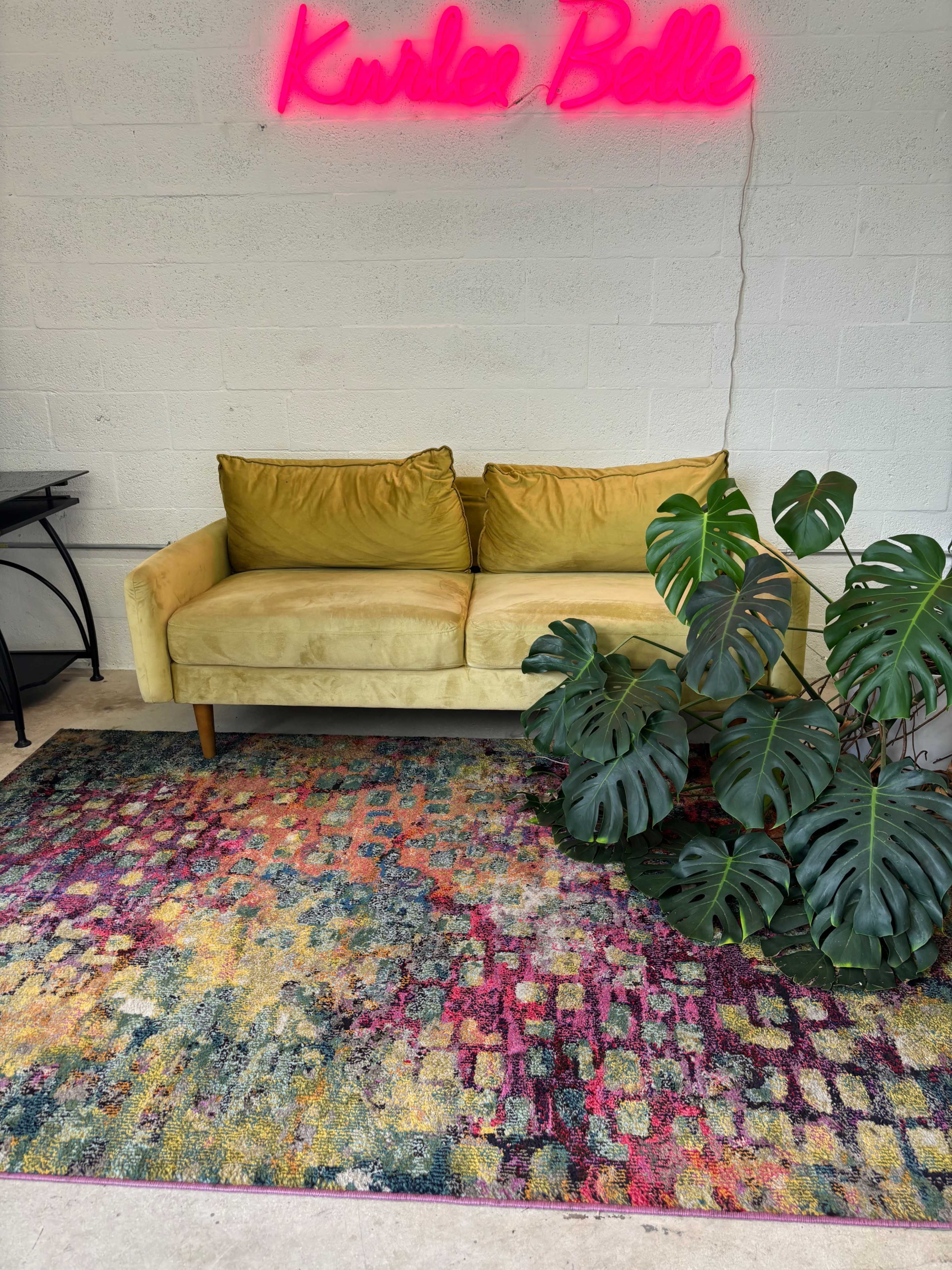 A yellow sofa sits against a white wall beneath a pink neon sign, accompanied by a large green plant and a colorful, patterned rug.
