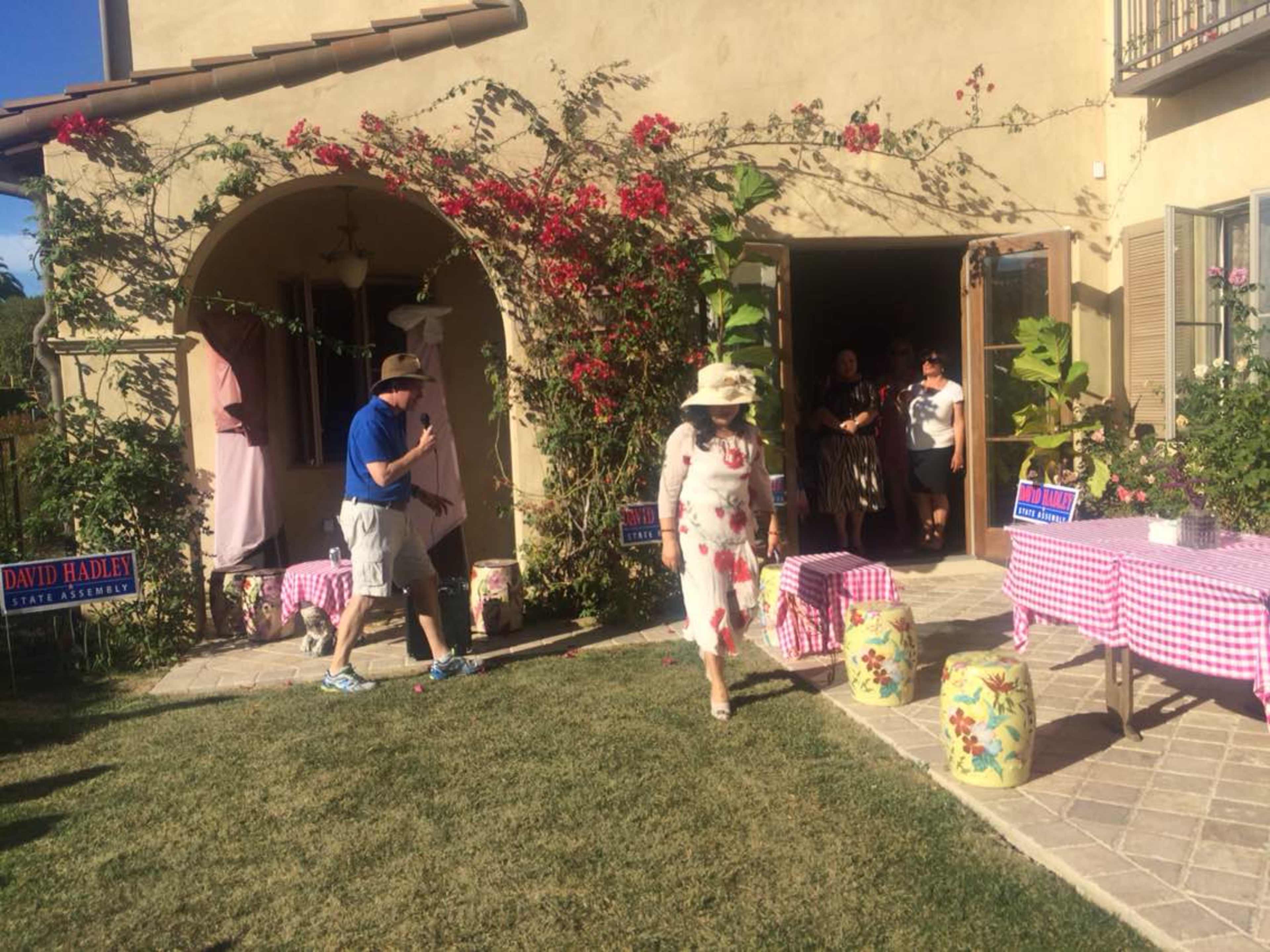 A group of people gathers outside a house adorned with flowers and campaign signs, while one individual in casual attire approaches from the left.