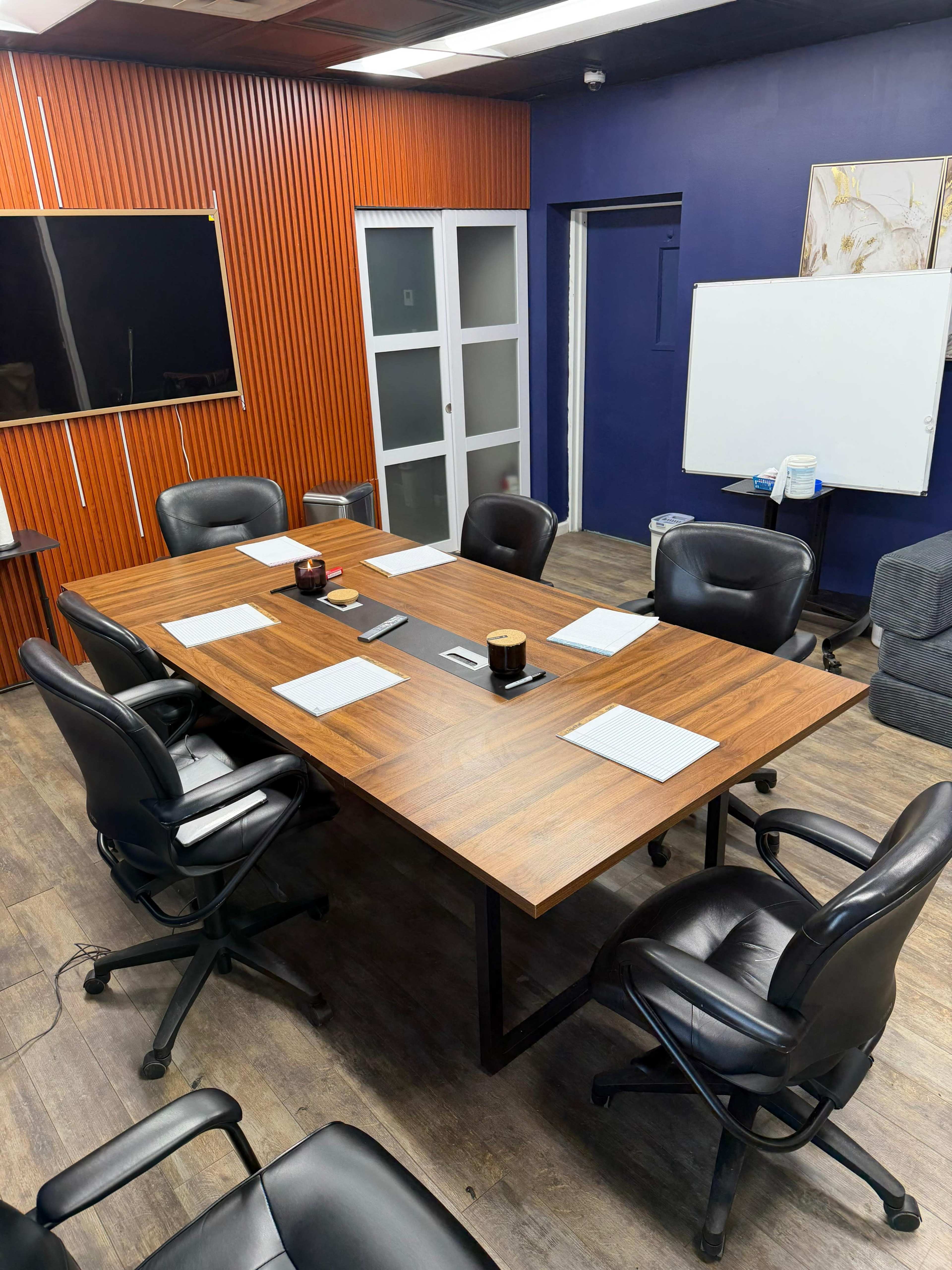 The image shows a conference room with a large wooden table, several black leather chairs, a whiteboard, and a presentation screen.