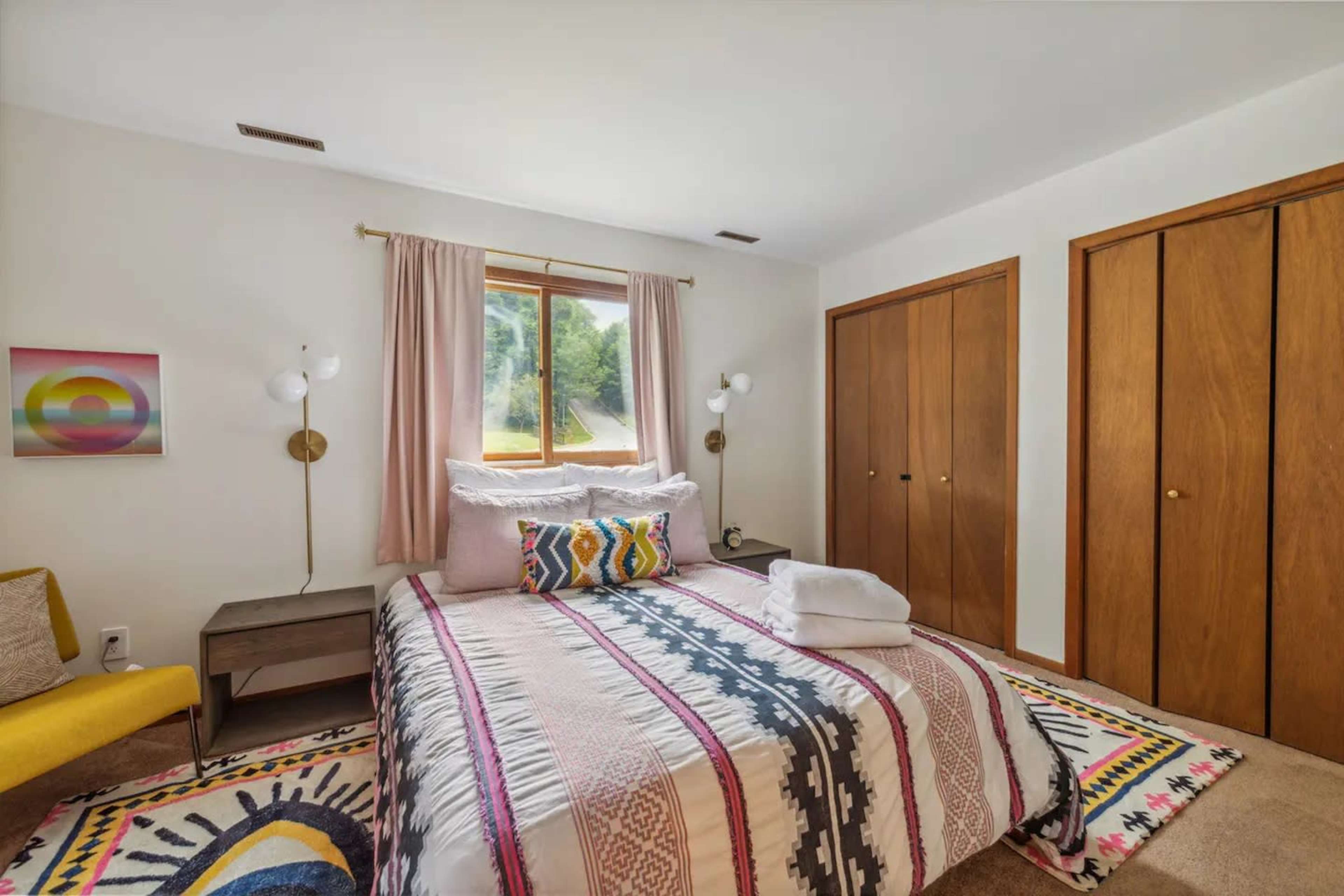 A cozy bedroom with a bed featuring a patterned blanket, two wooden closet doors, and a window allowing natural light to enter.