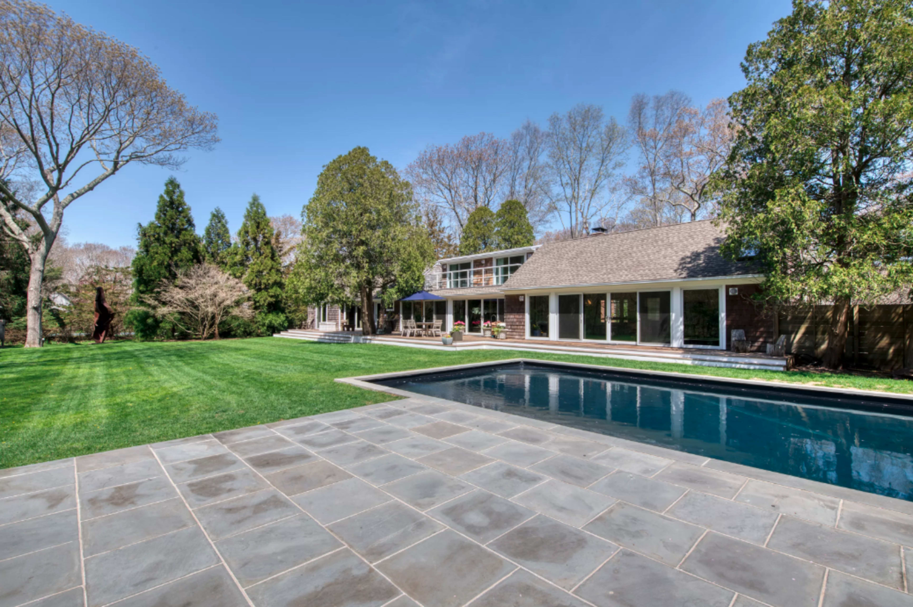 A modern house with a stone patio and a swimming pool, surrounded by a well-kept lawn and trees in the background.