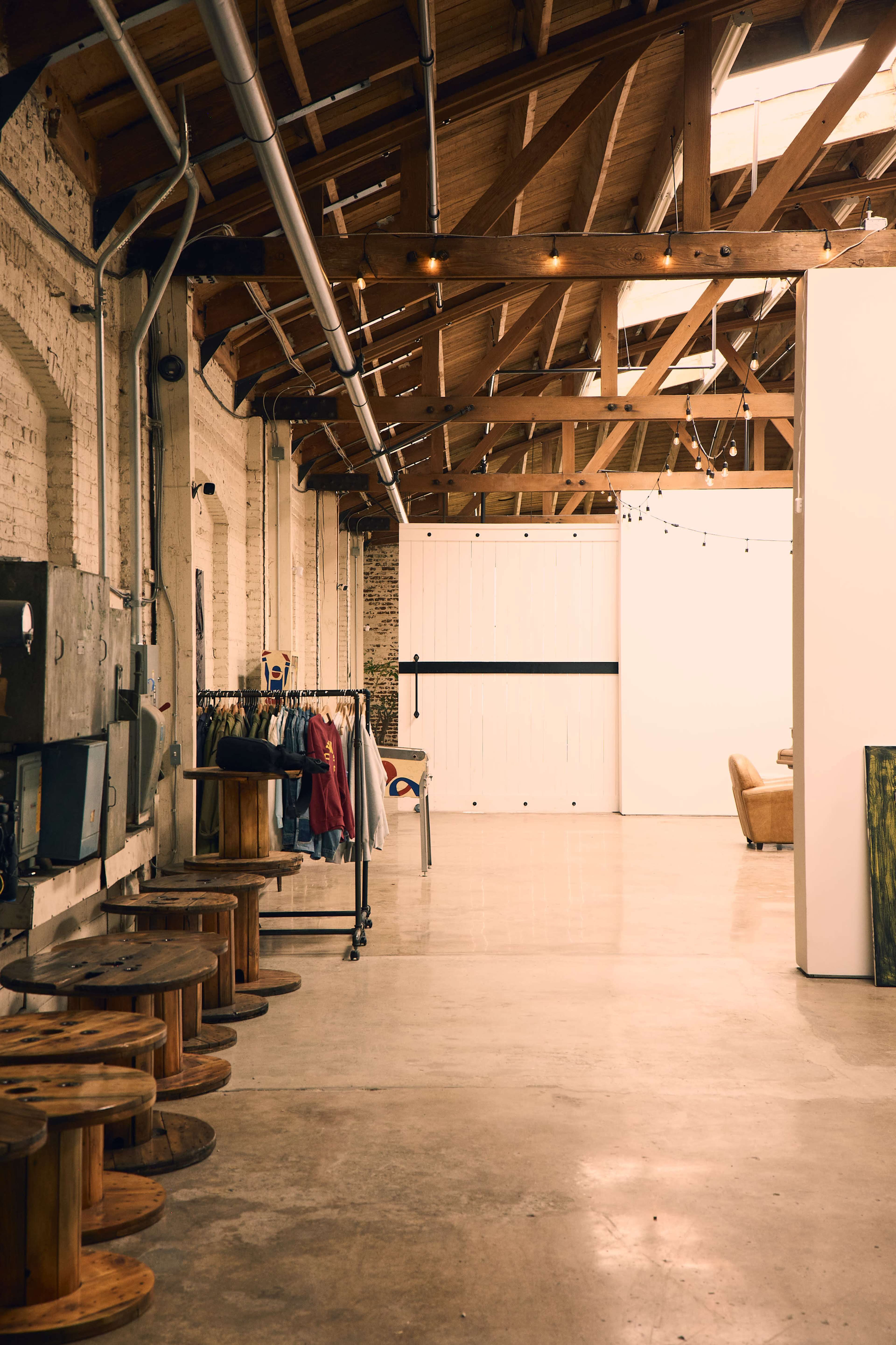 The image shows an industrial-style interior with a lofted wooden ceiling, clothing racks on one side, and a large white partition in the background.