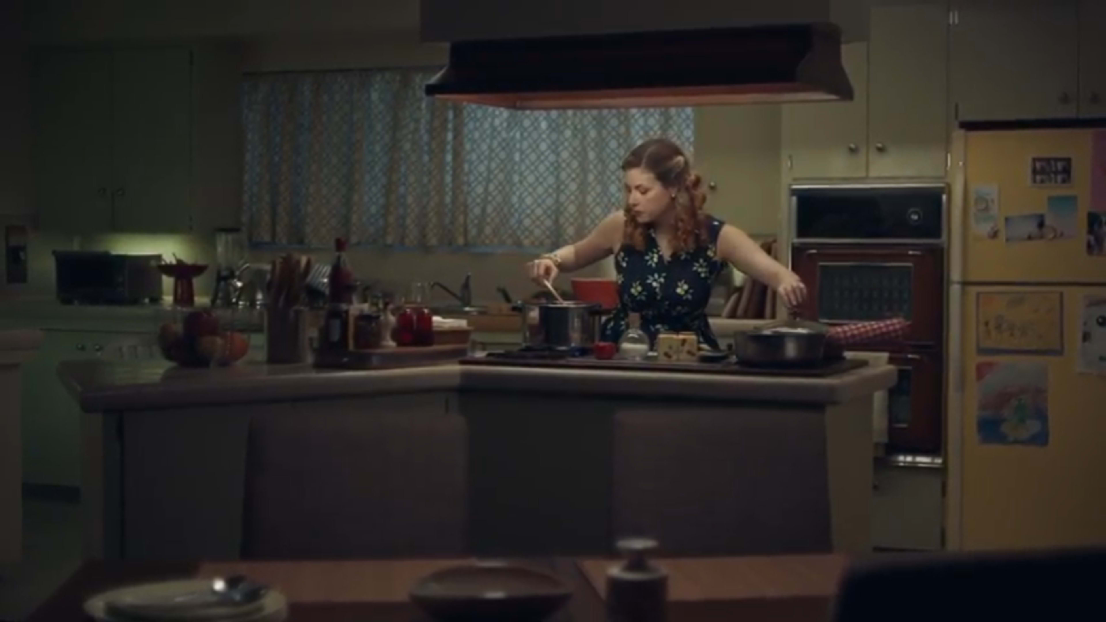 A woman cooks at a kitchen island while several pots are arranged on the stove, with a colorful fridge and scattered kitchen items in the background.