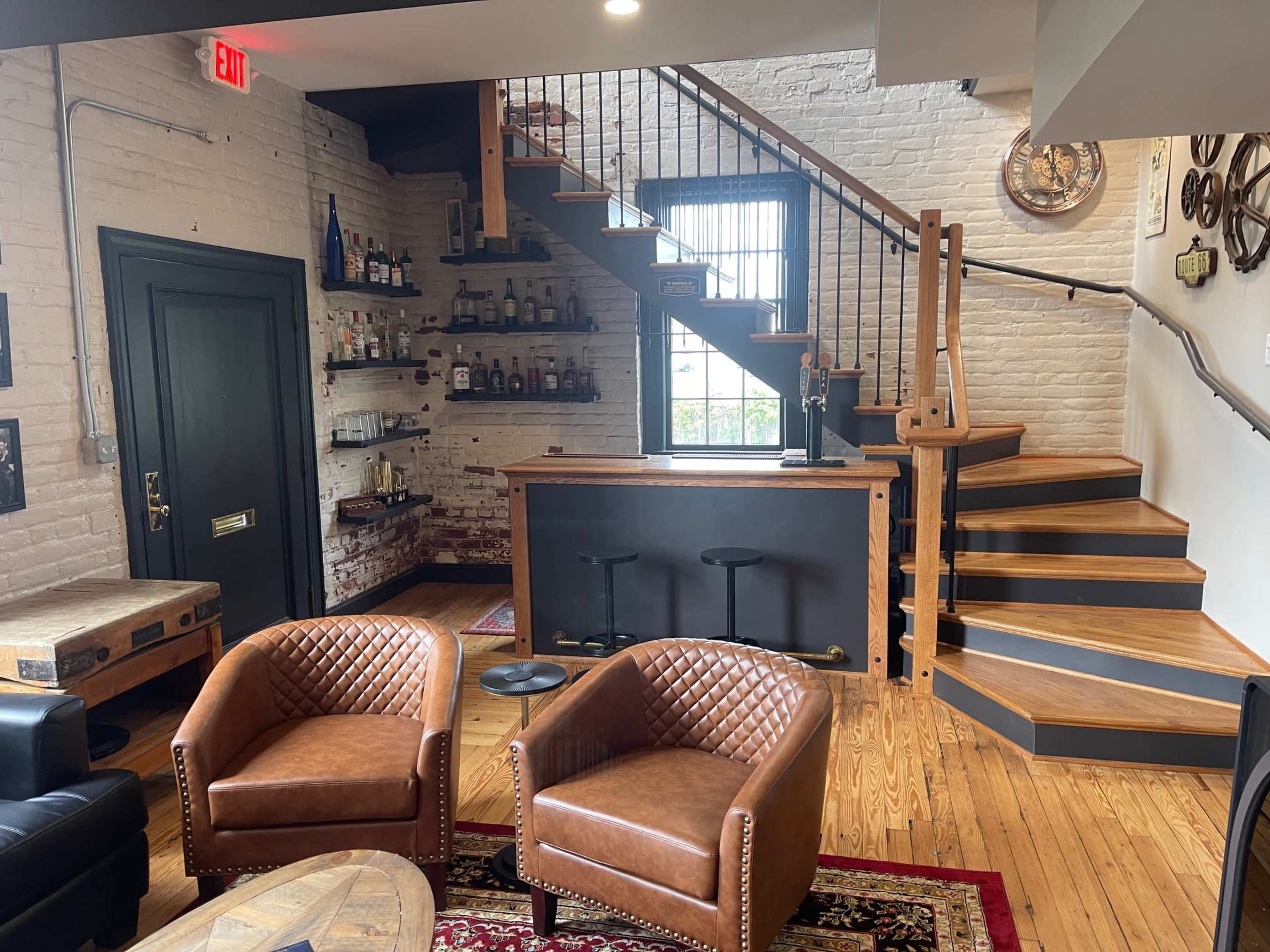 The image shows a cozy interior space featuring a bar area on the left, a staircase leading to an upper level, and two brown leather chairs arranged on a wooden floor.