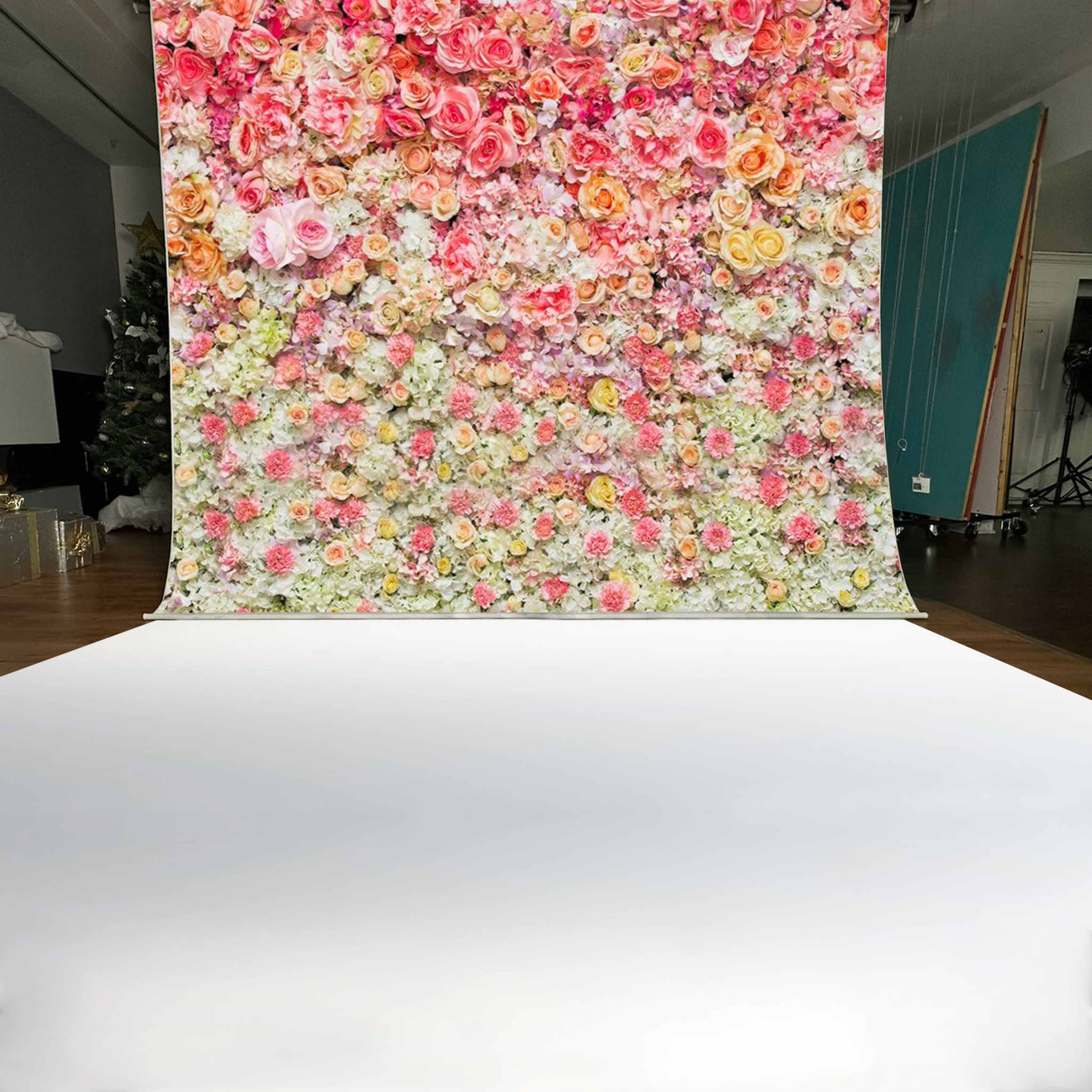 The image features a backdrop covered with an arrangement of colorful artificial flowers in various shades, set against a plain white floor.