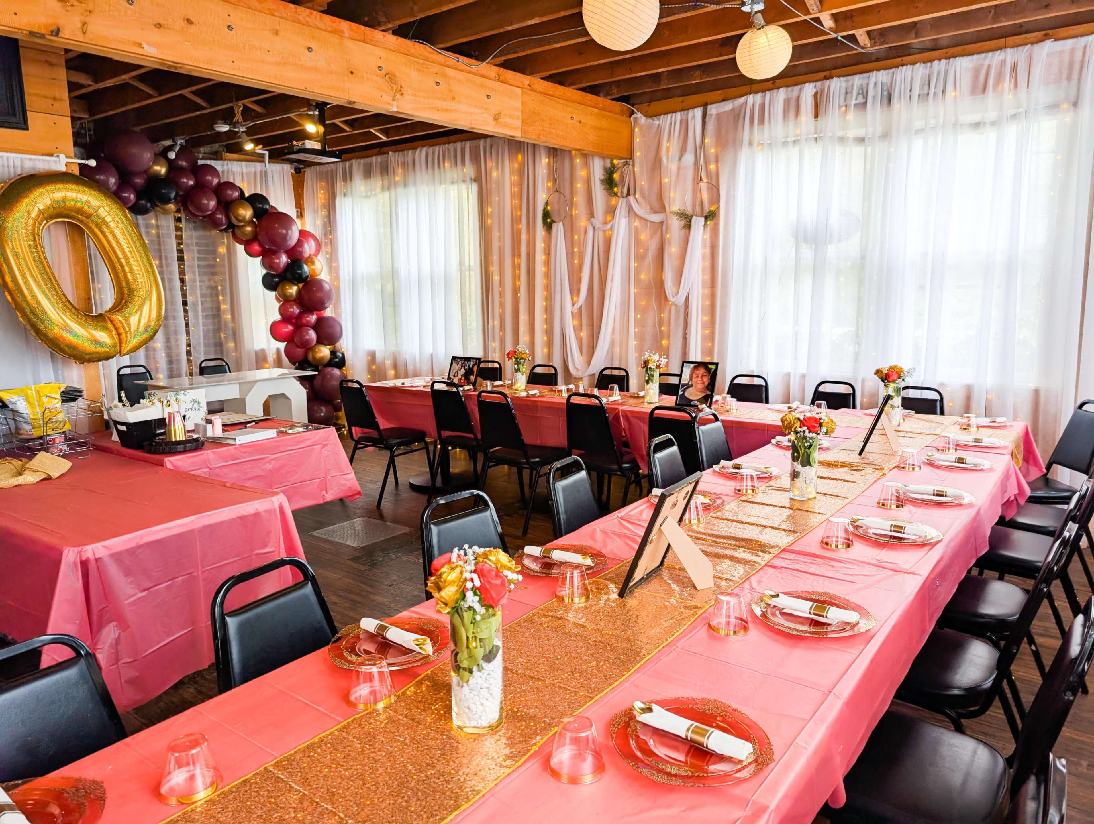 The image shows a decorated event space with tables covered in pink tablecloths, a long glittery centerpiece, balloons in various colors, and floral arrangements.