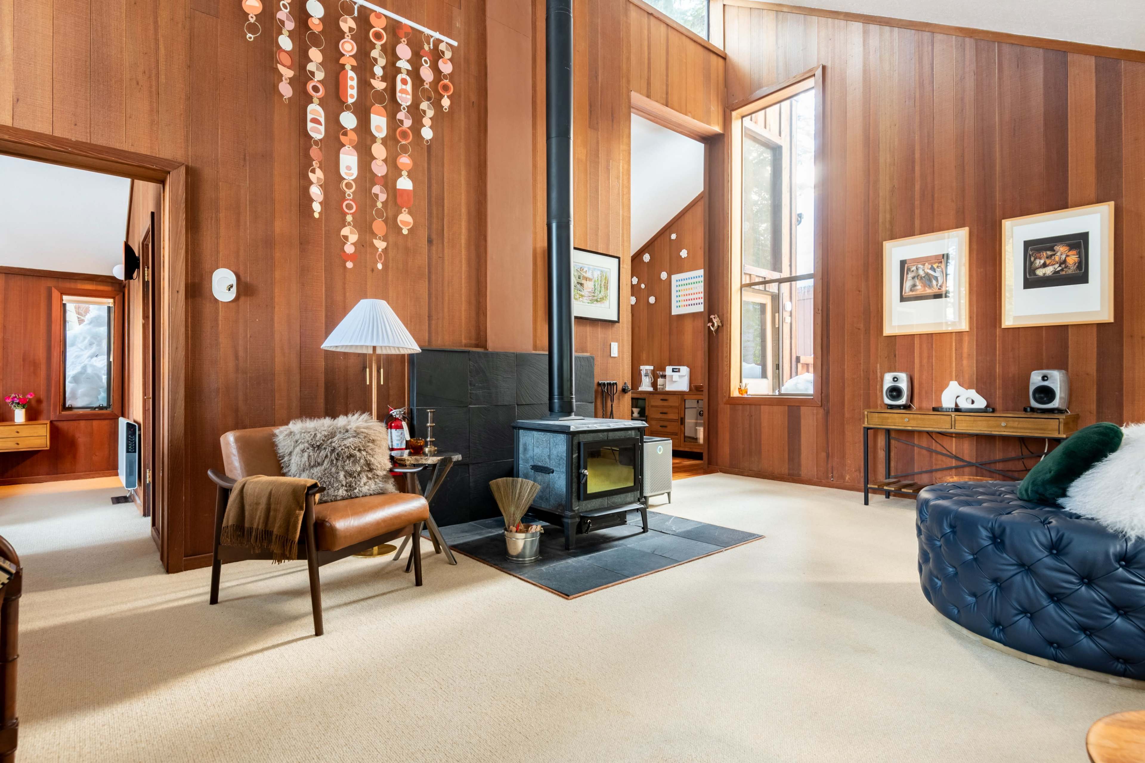 A wooden-paneled living room with a wood stove, a cozy armchair, and large windows allowing natural light to enter.
