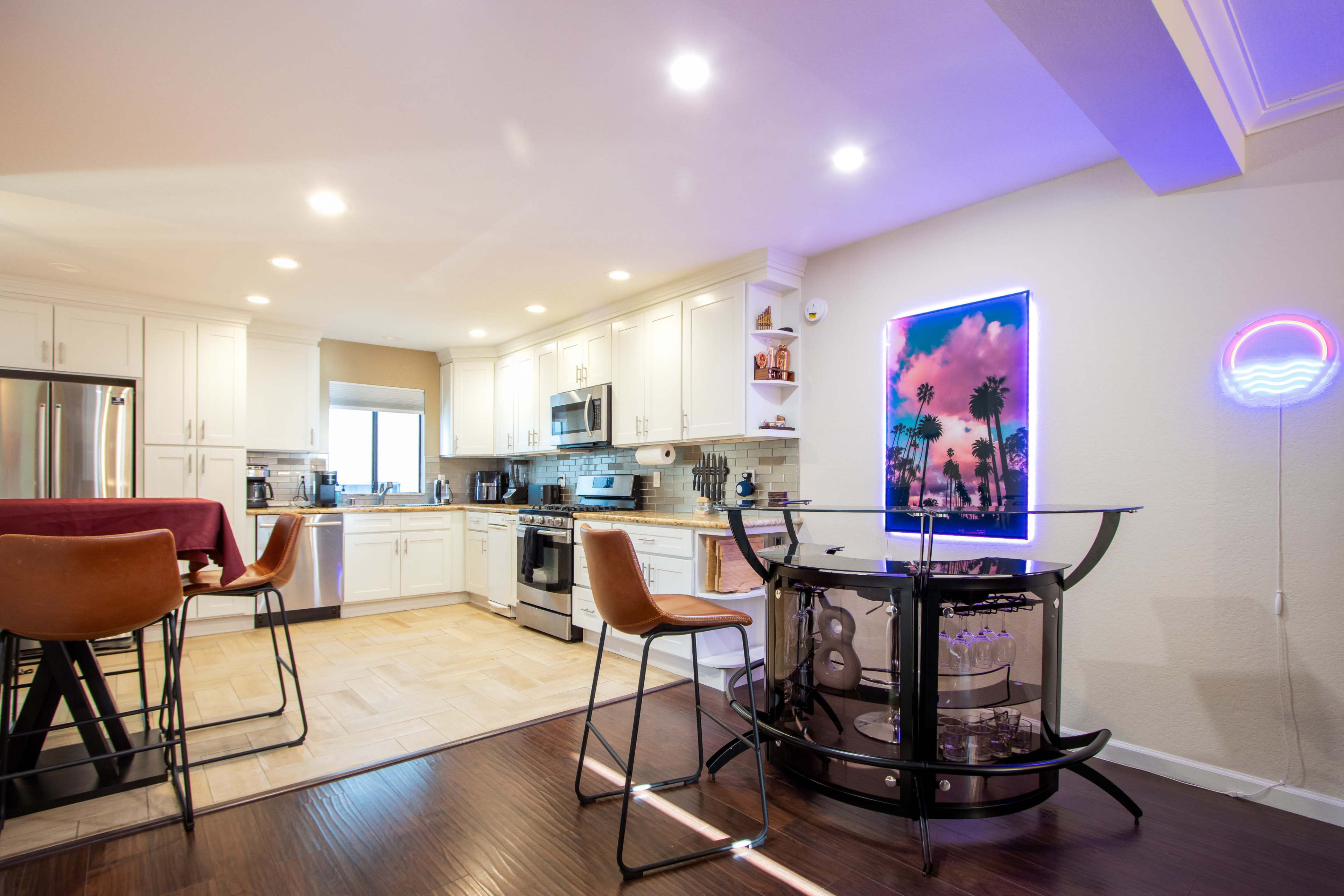 A modern kitchen with white cabinets, stainless steel appliances, and a bar area featuring two stools and a neon sign.