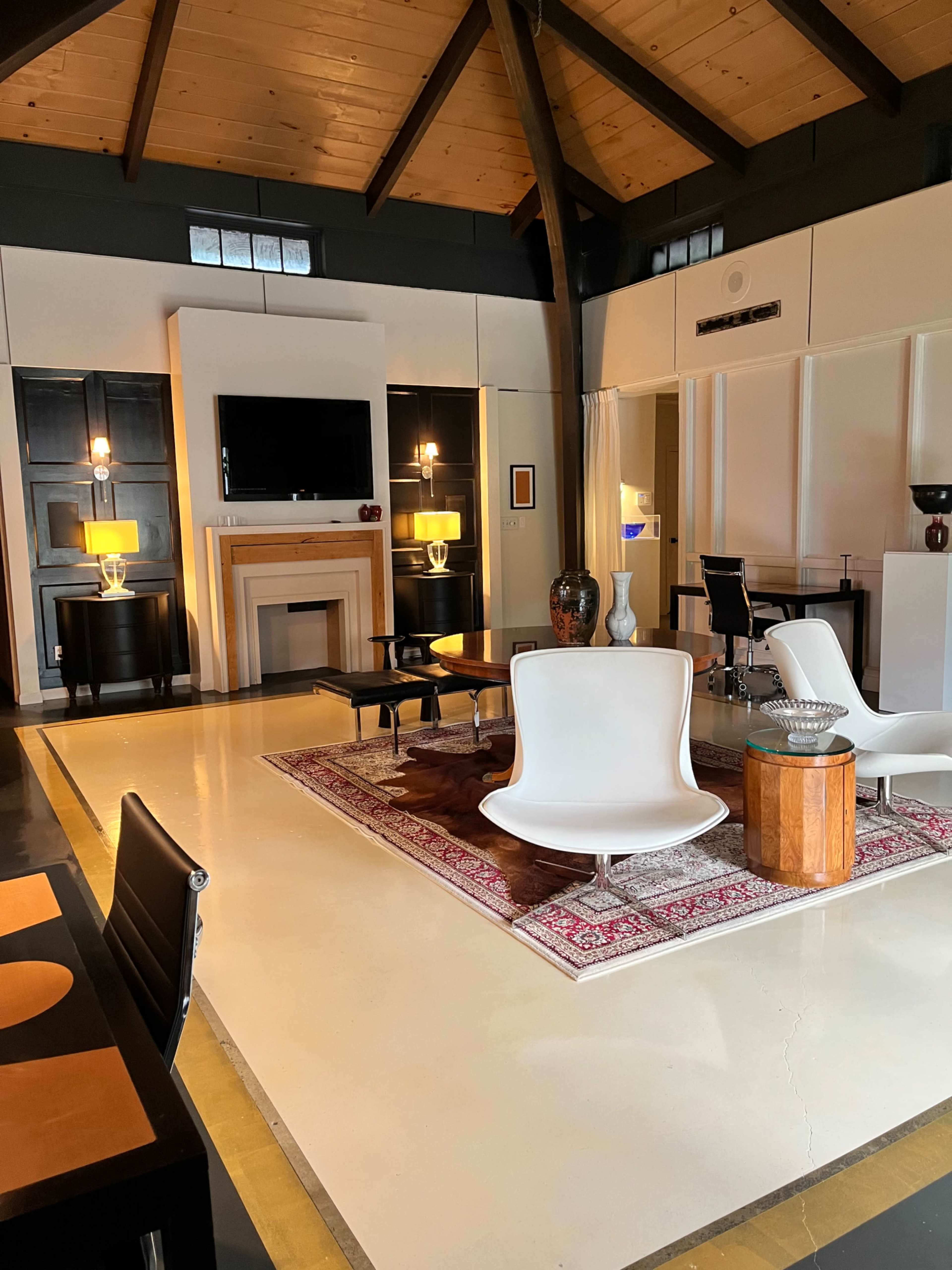 The image shows a spacious living area with wooden beams, modern furniture, a large television, and decorative elements like a rug and vases.