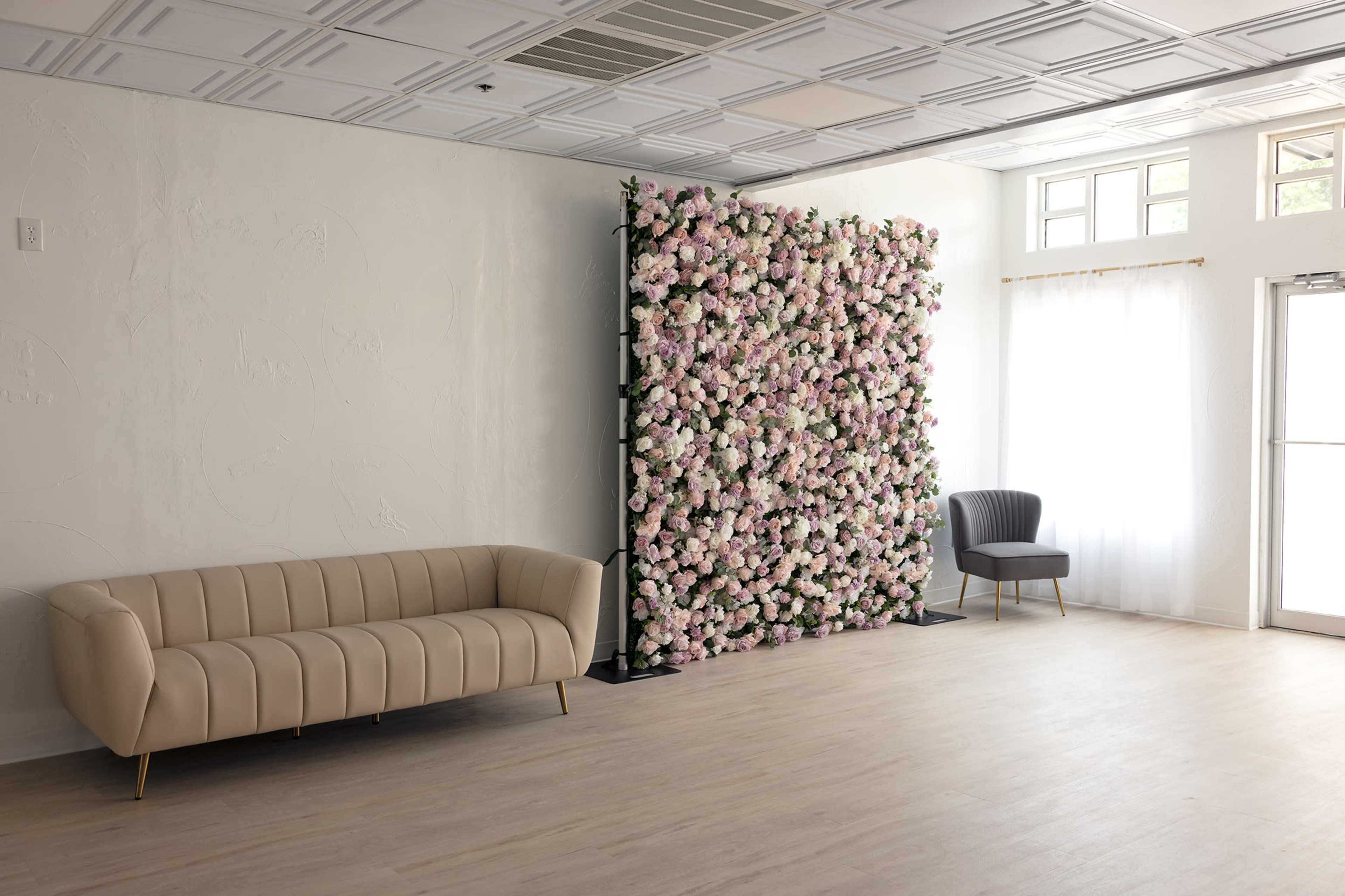 The image features a spacious room with a beige sofa and a gray armchair, complemented by a floral wall decoration made up of pink and white flowers.