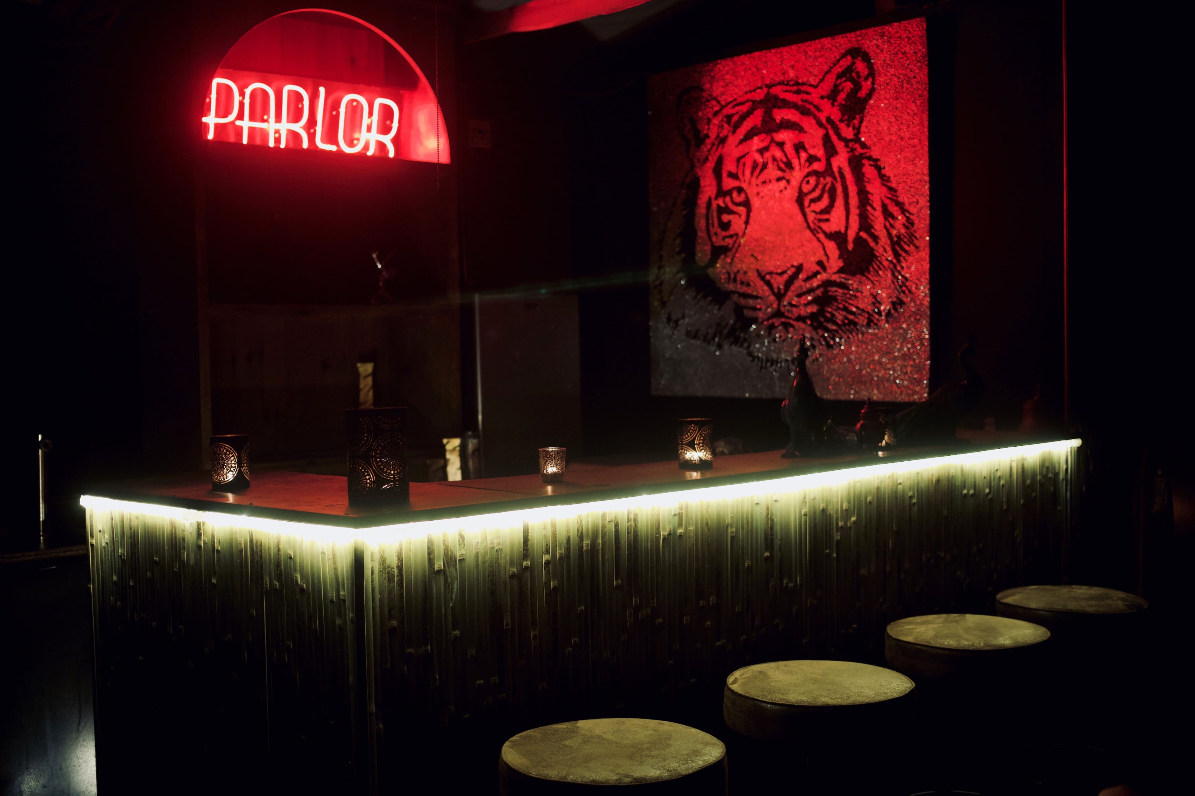 A dimly lit bar with a neon "PARLOR" sign and a large tiger portrait on the wall features wooden stools and candles on the counter.