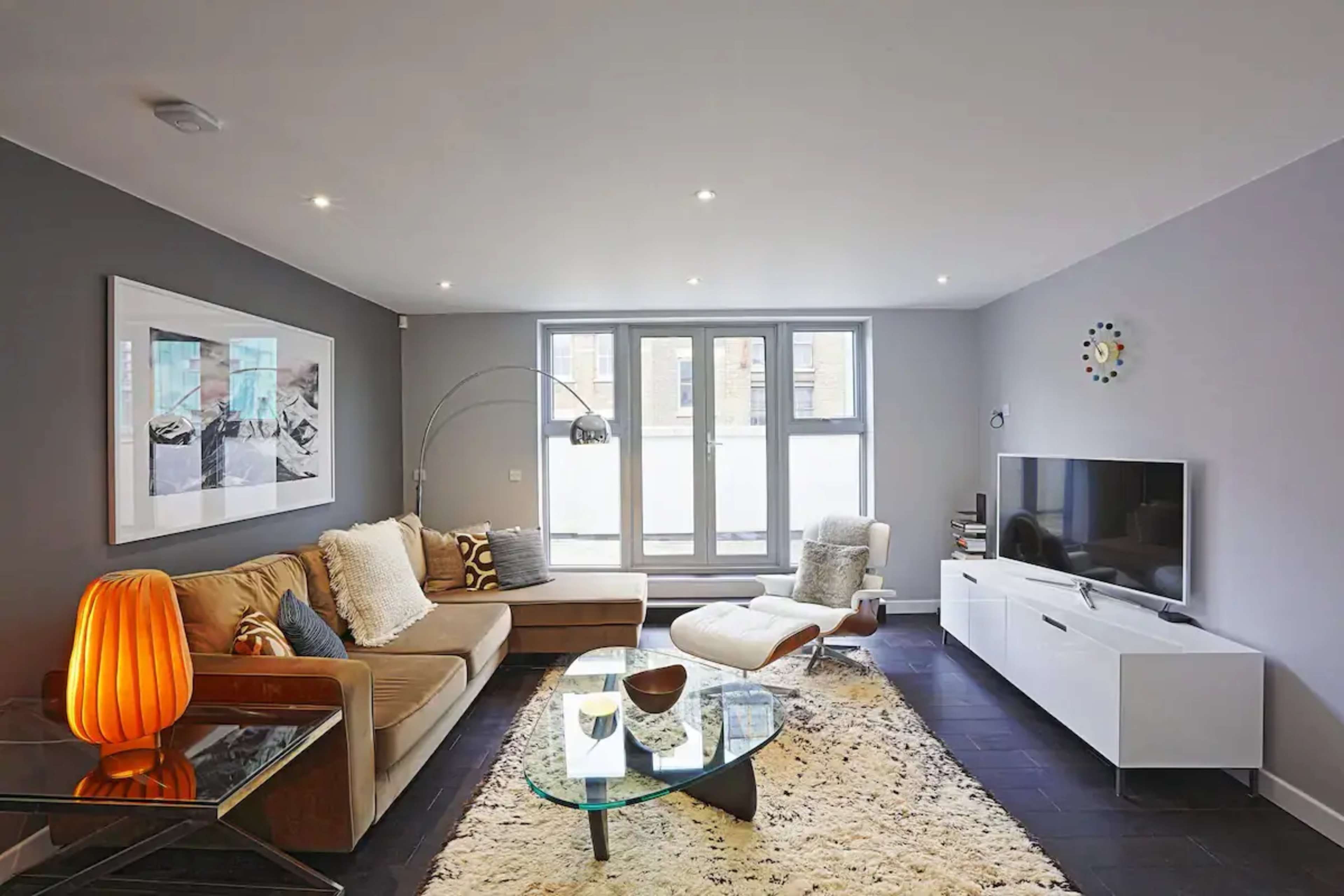 A modern living room features a sectional sofa, a glass coffee table, and a television on a white cabinet, illuminated by ceiling lights and natural light from large windows.