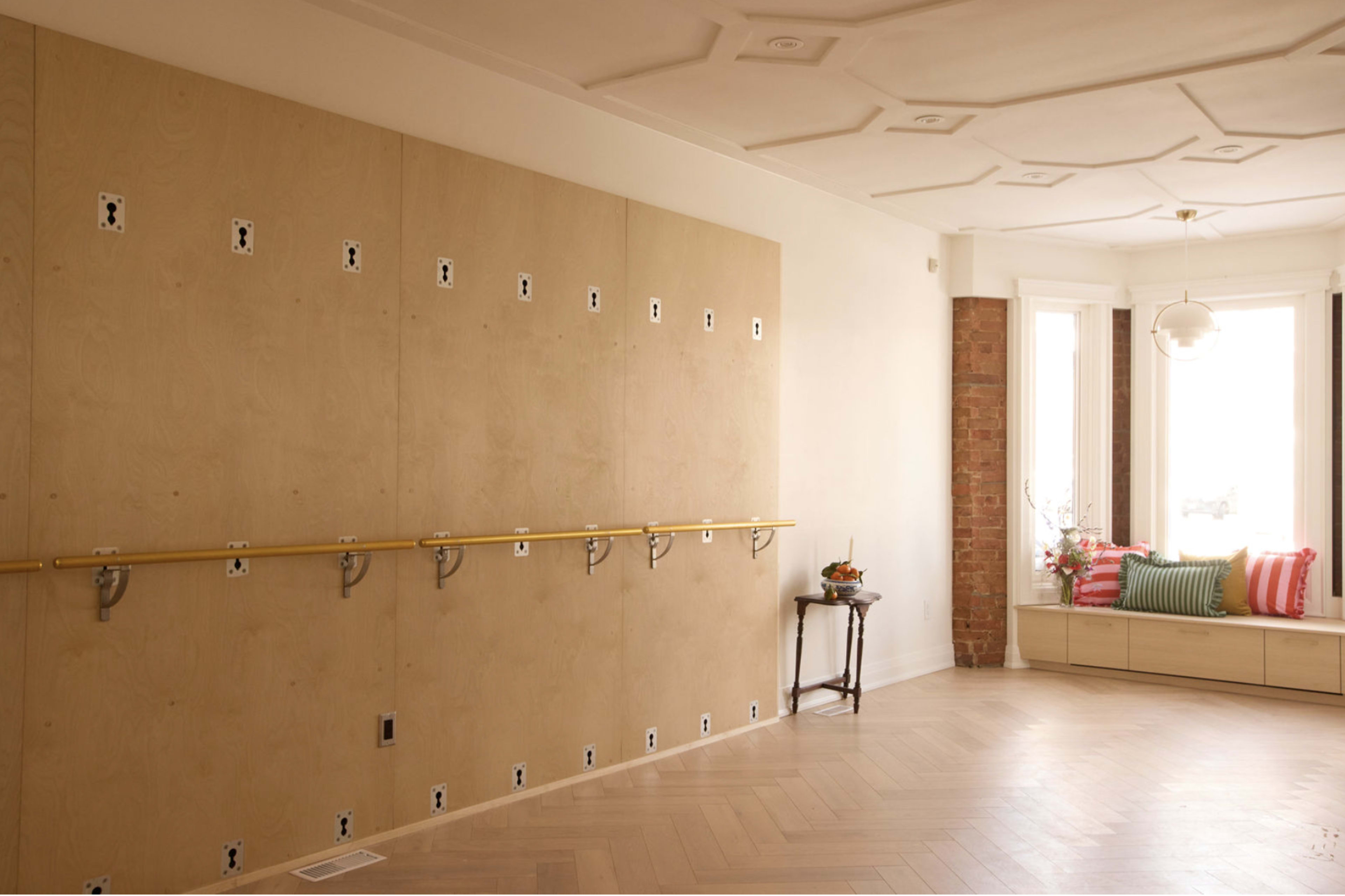 The image shows a spacious room with a wooden floor, featuring a wall equipped with multiple electrical outlets and a ballet barre mounted along its length, alongside a bright window nook with cushions.