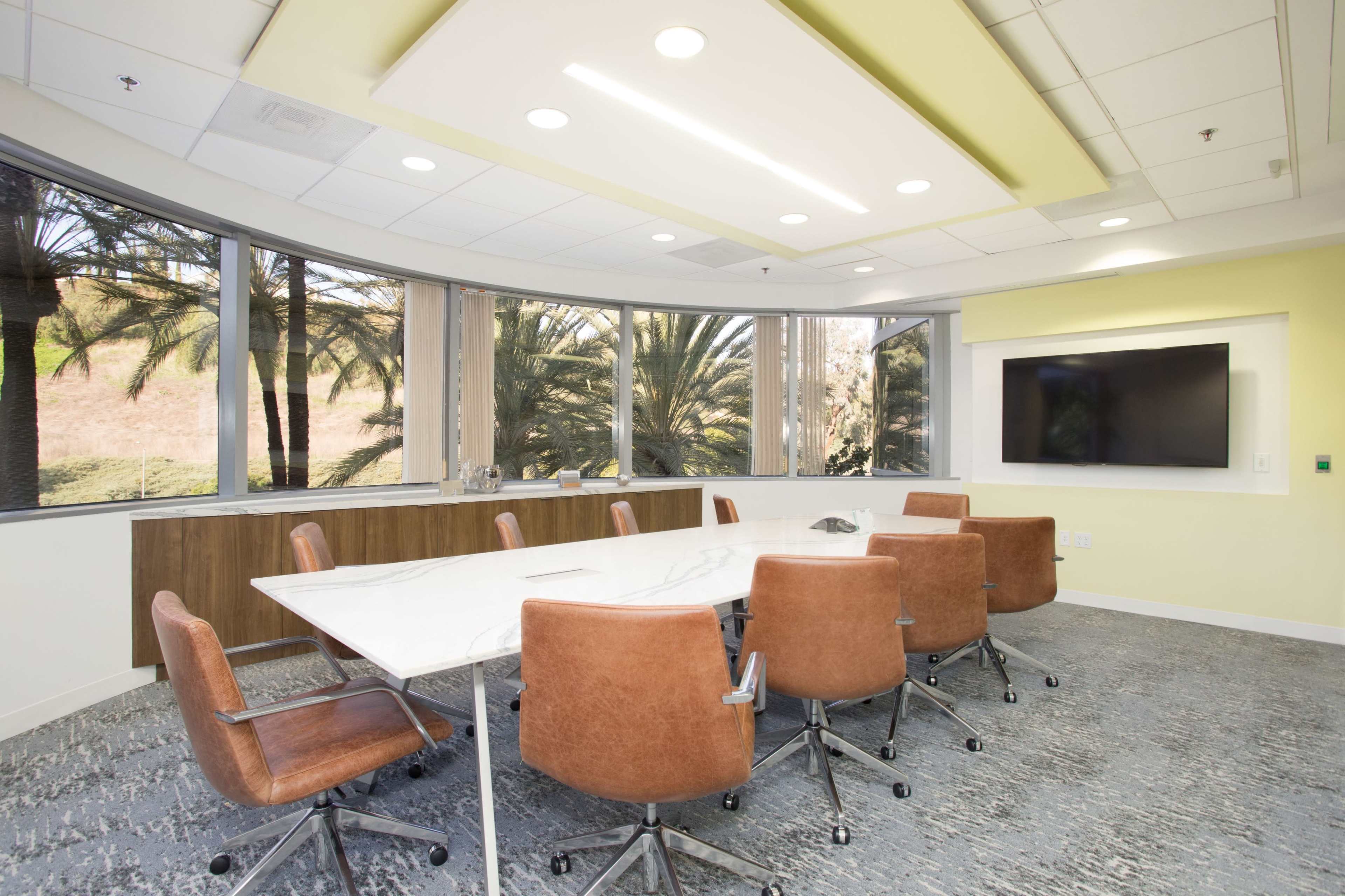 A modern conference room features a large white table surrounded by eight brown chairs, with large windows overlooking palm trees.