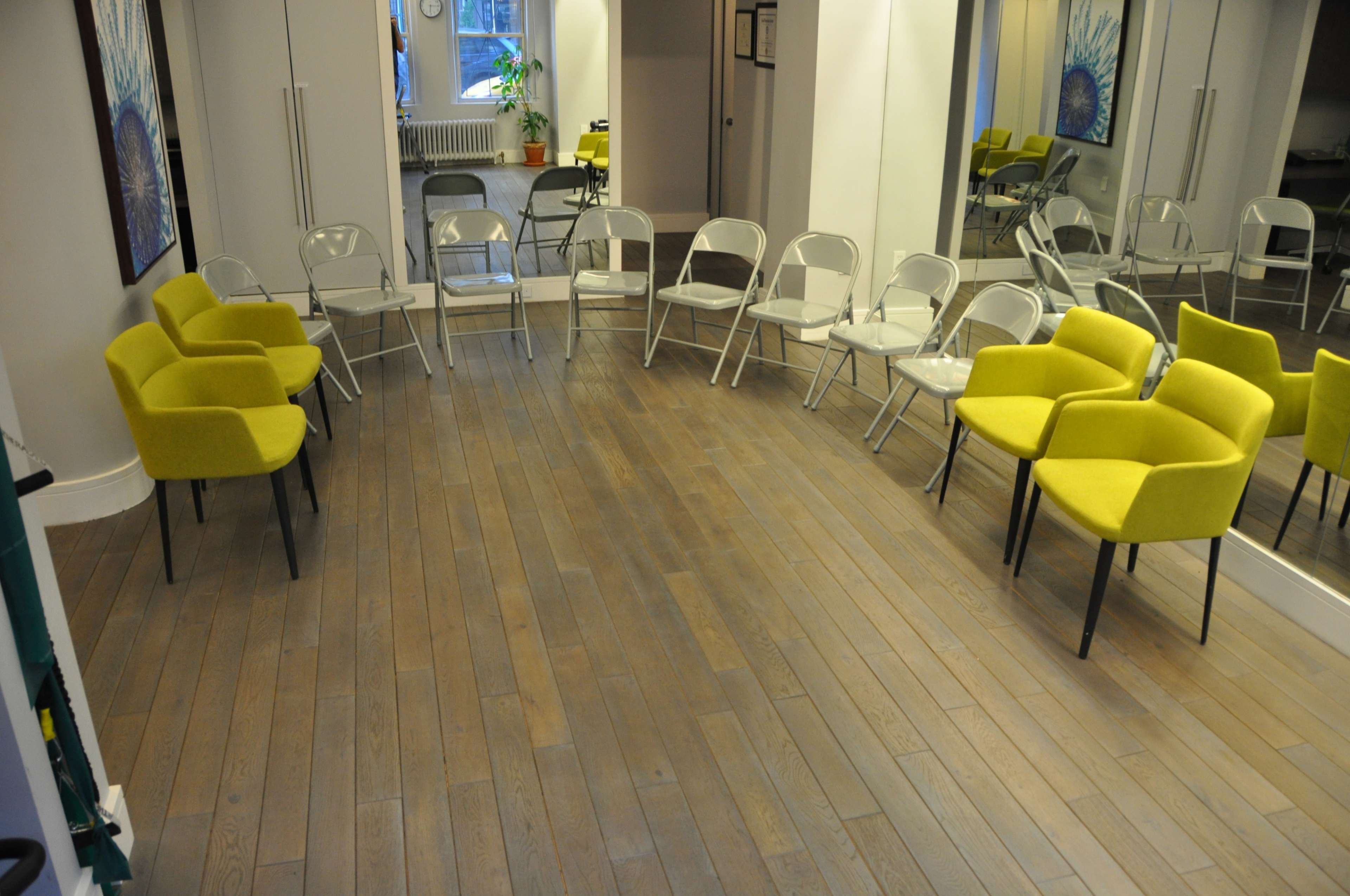 A spacious room with wooden flooring, featuring yellow armchairs and transparent folding chairs arranged in a circular setup around mirrored walls.