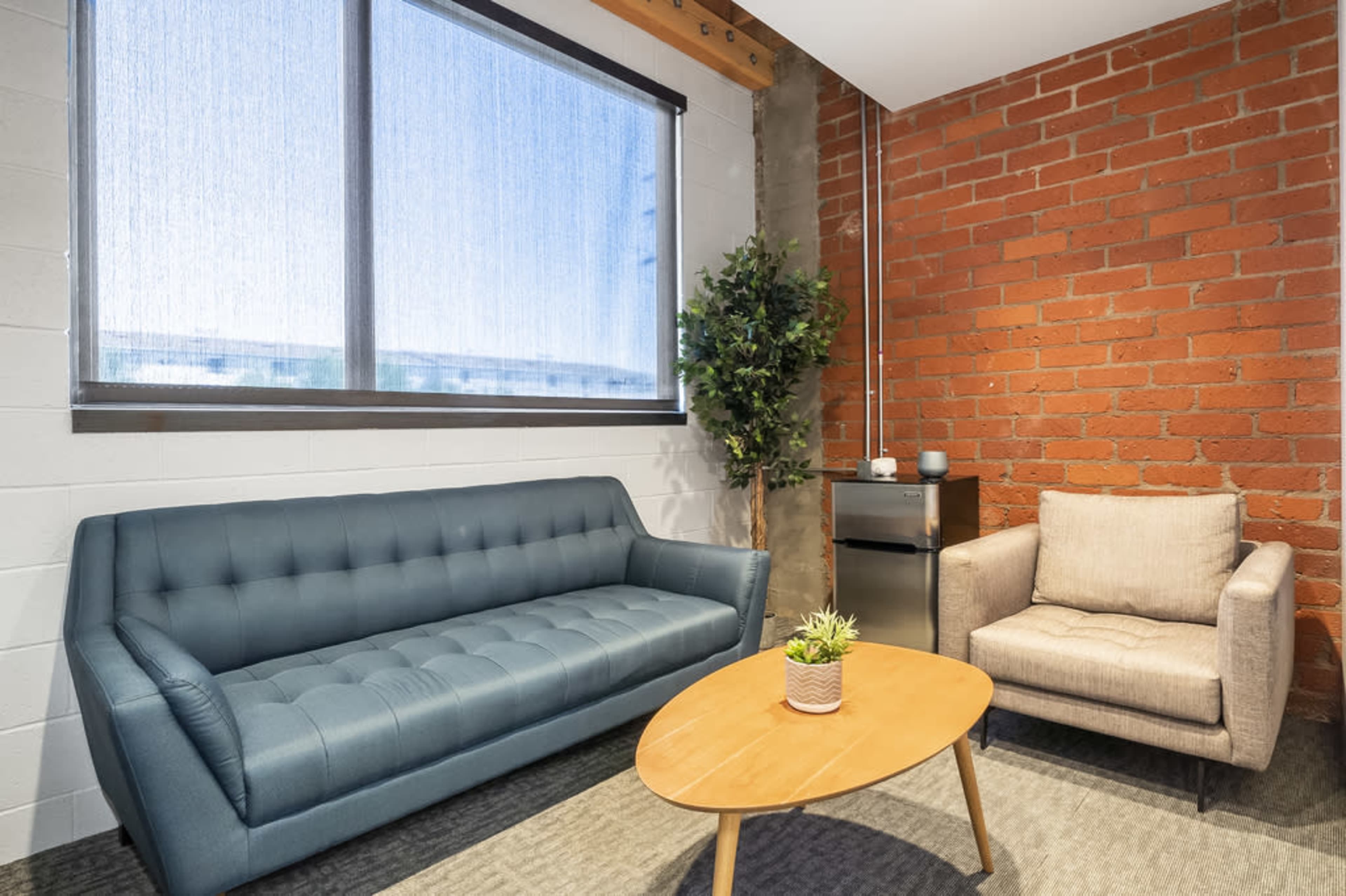 The image shows a small modern lounge area featuring a blue sofa, a beige armchair, a wooden coffee table, and a potted plant, with a large window allowing natural light to enter.