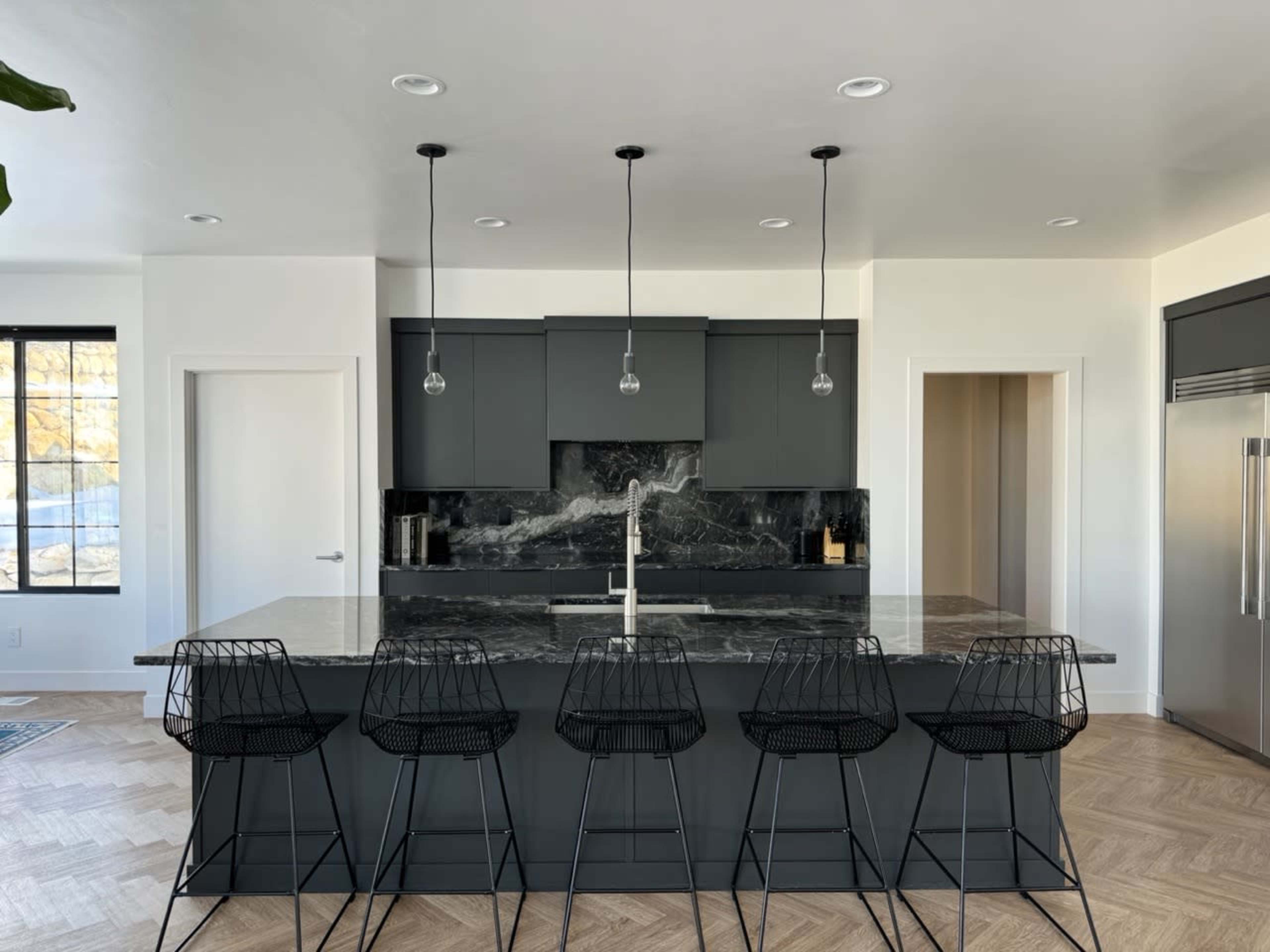 A modern kitchen features a large island with a dark marble countertop and five black wire bar stools.