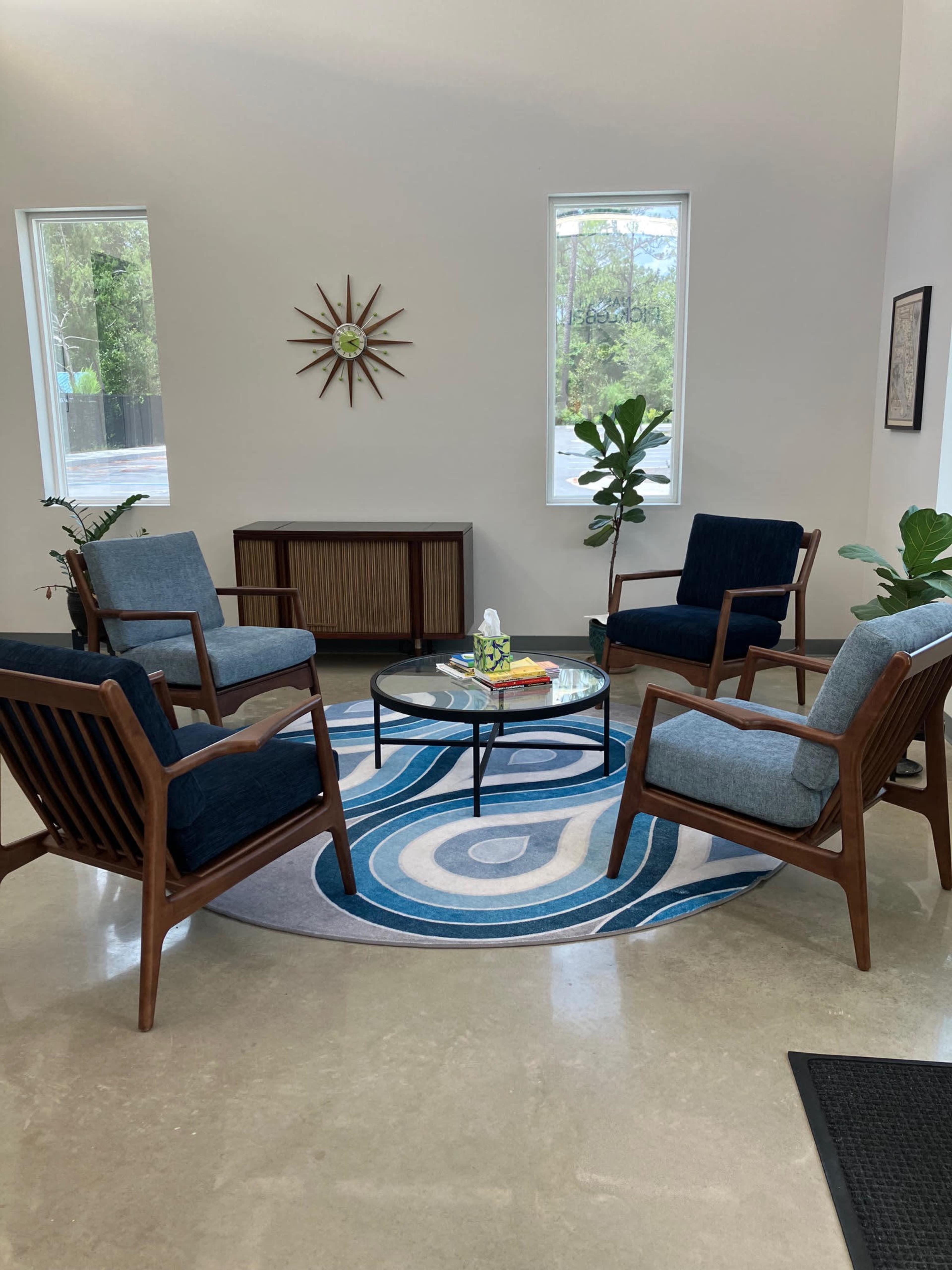 The image shows a modern seating area with four wooden chairs arranged around a circular glass coffee table, set on a patterned rug, with plants and windows in the background.