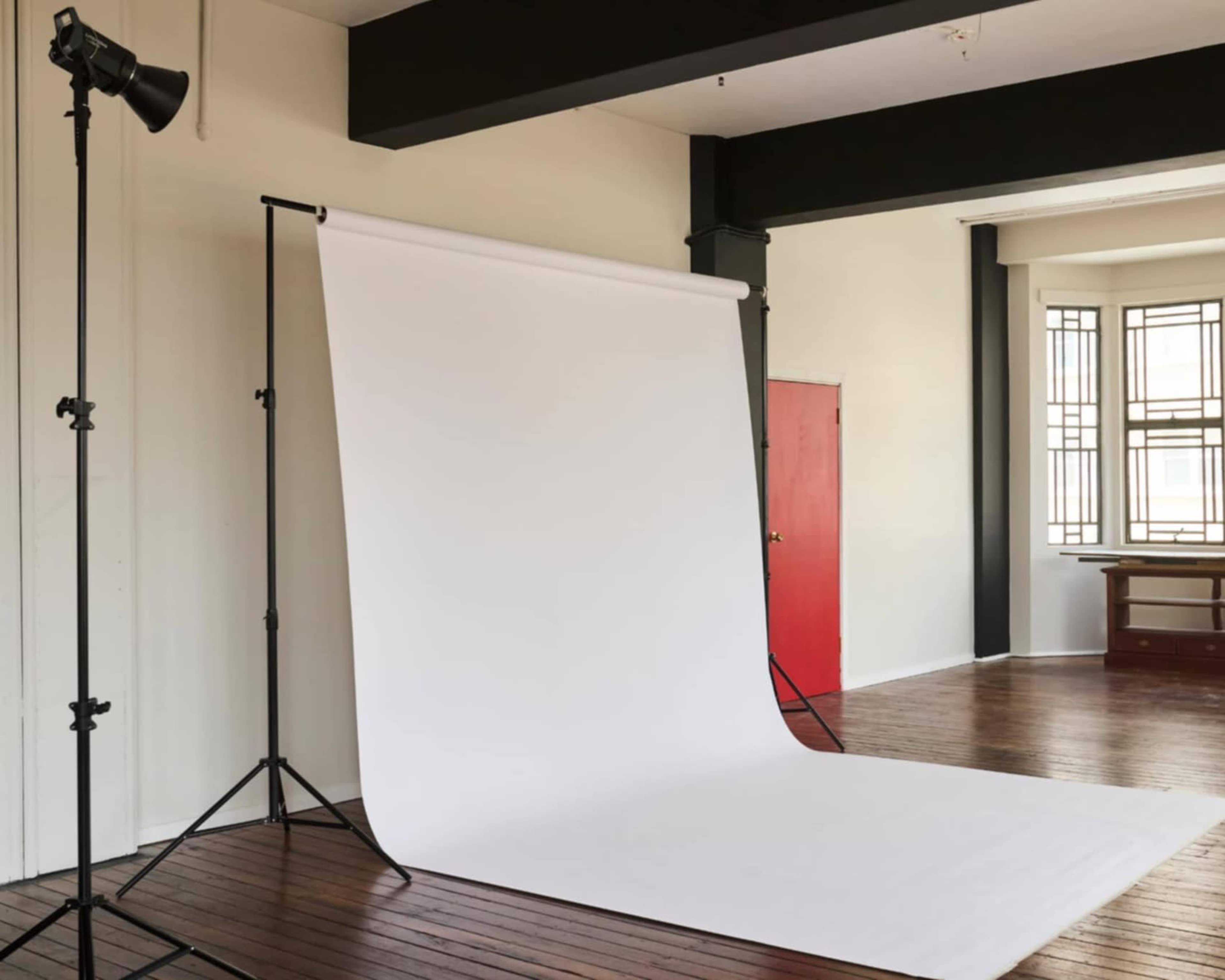 The image shows a photography studio featuring a white backdrop on a stand, with a light source positioned nearby and a red door visible in the background.