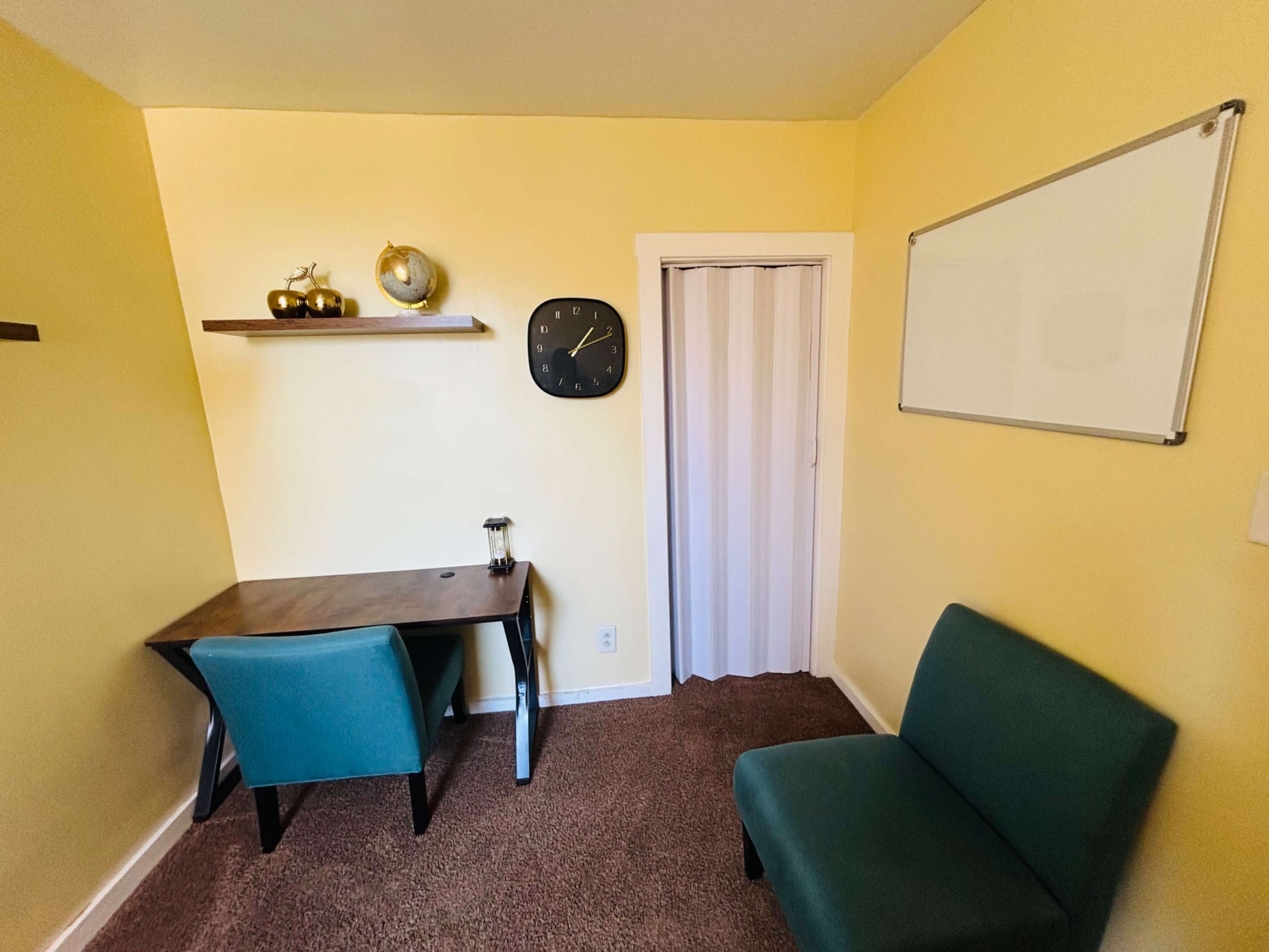 The image shows a small, yellow room with a wooden desk, a clock on the wall, a whiteboard, and two teal chairs.