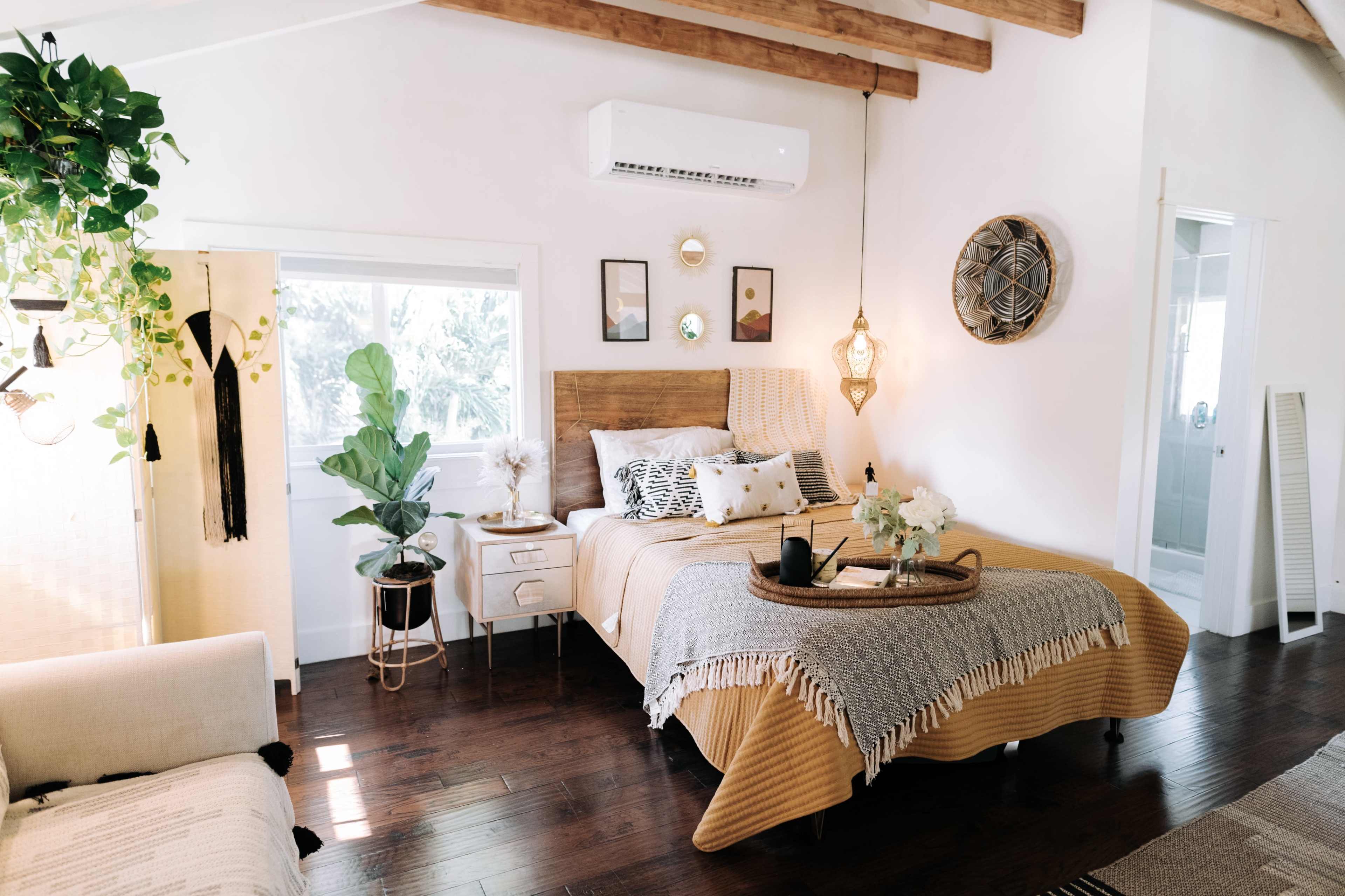 A cozy bedroom features a large bed with a gold blanket, potted plants, and decorative wall art beneath wooden beams and an air conditioning unit.