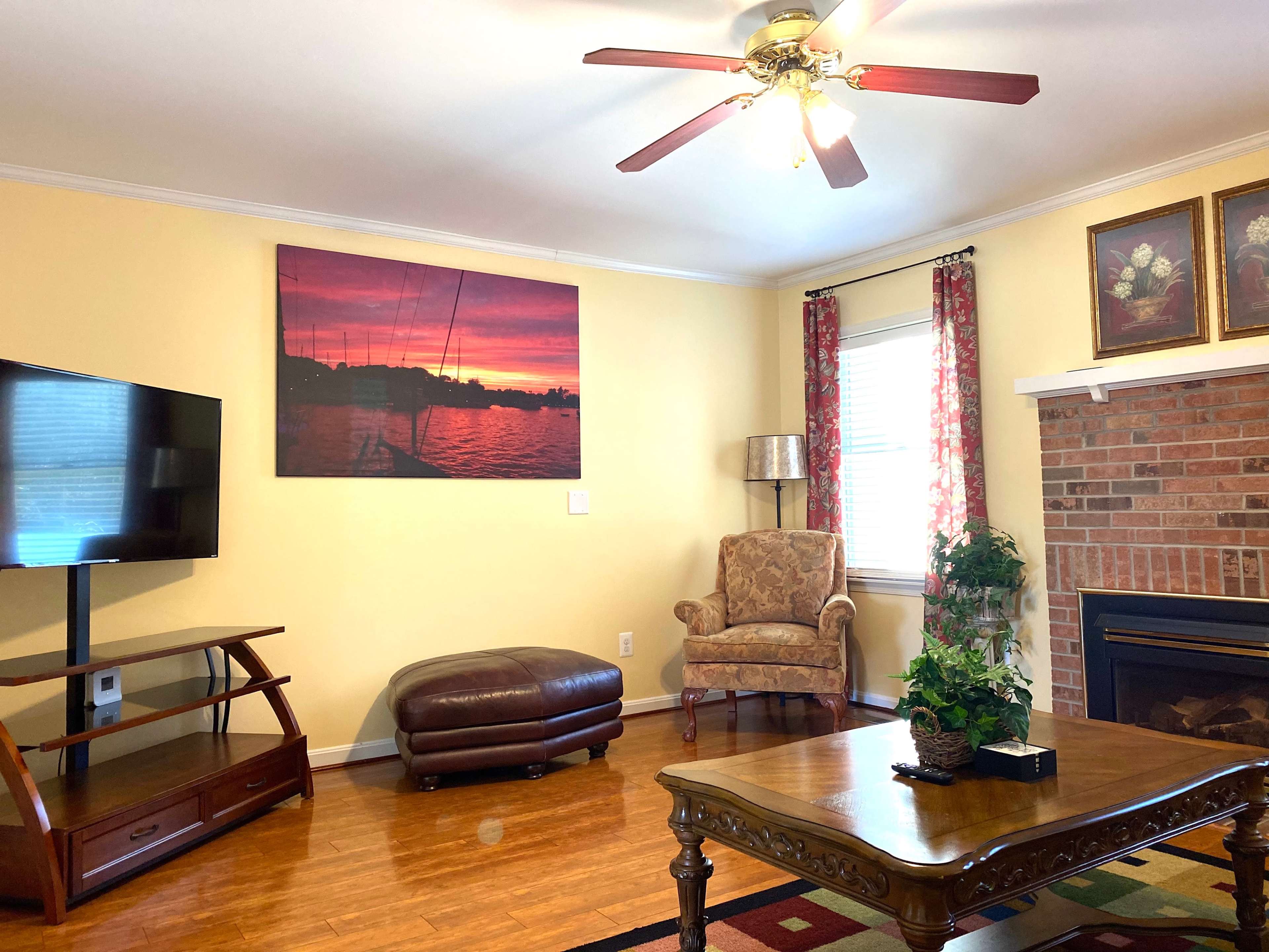 The living room features a television on a stand, a brown ottoman, a patterned rug, and a chair beside a fireplace, with a large sunset painting on the wall.