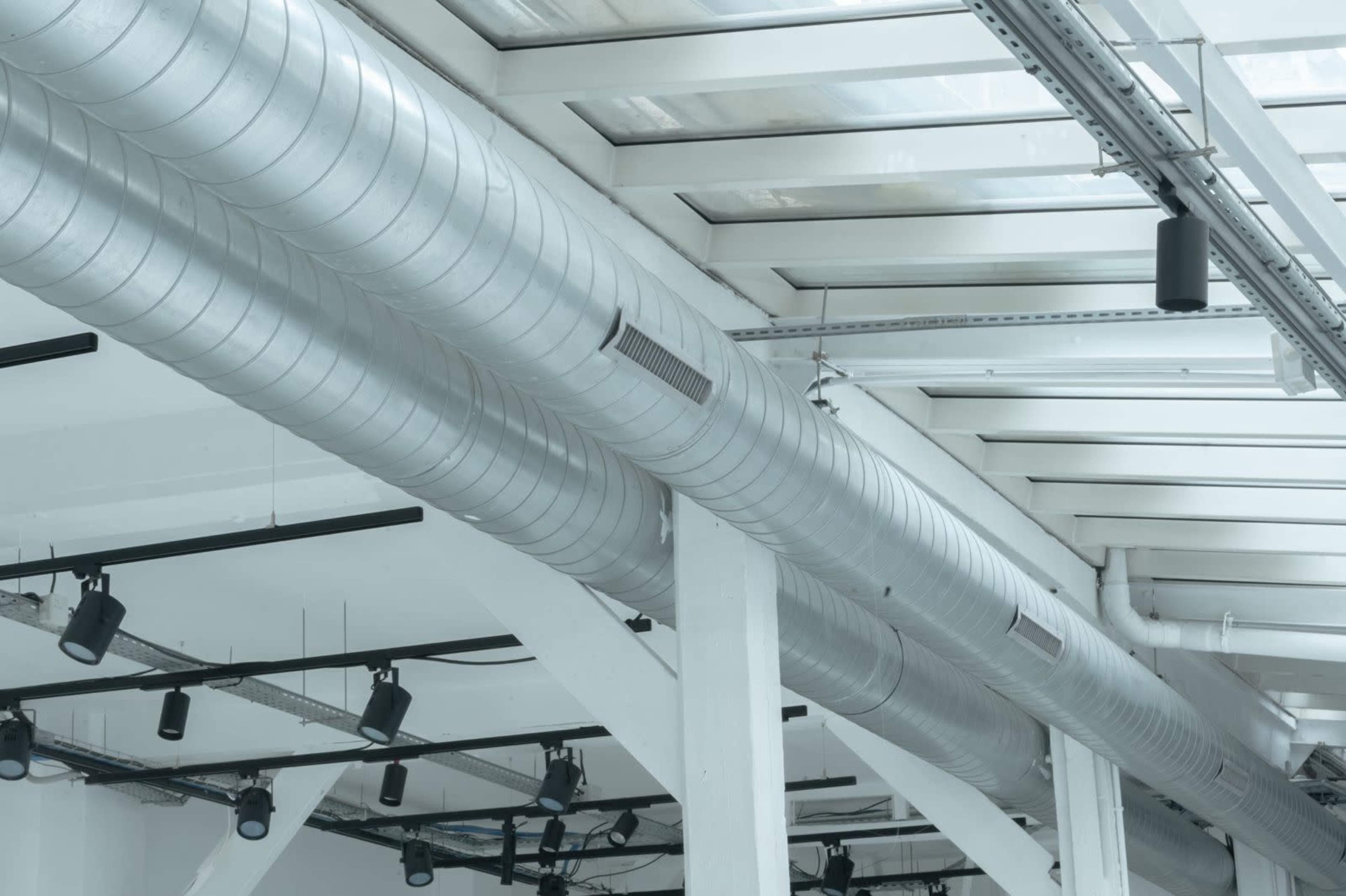 The image shows large metallic ductwork running along a modern building's ceiling with exposed beams and overhead lighting.
