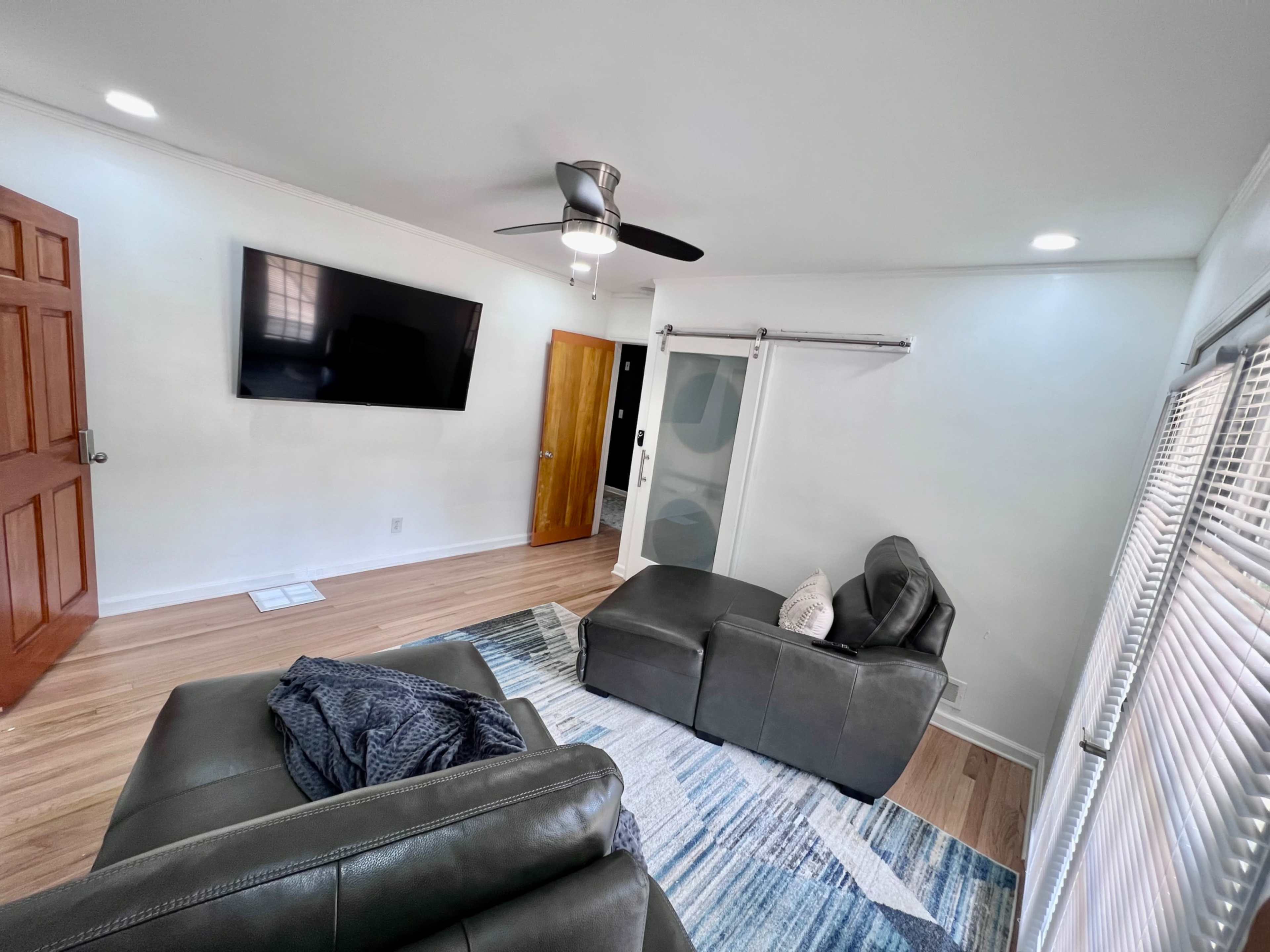 The living room features a gray leather sofa and armchair, a mounted television on the wall, and a sliding door with a wooden frame next to it.