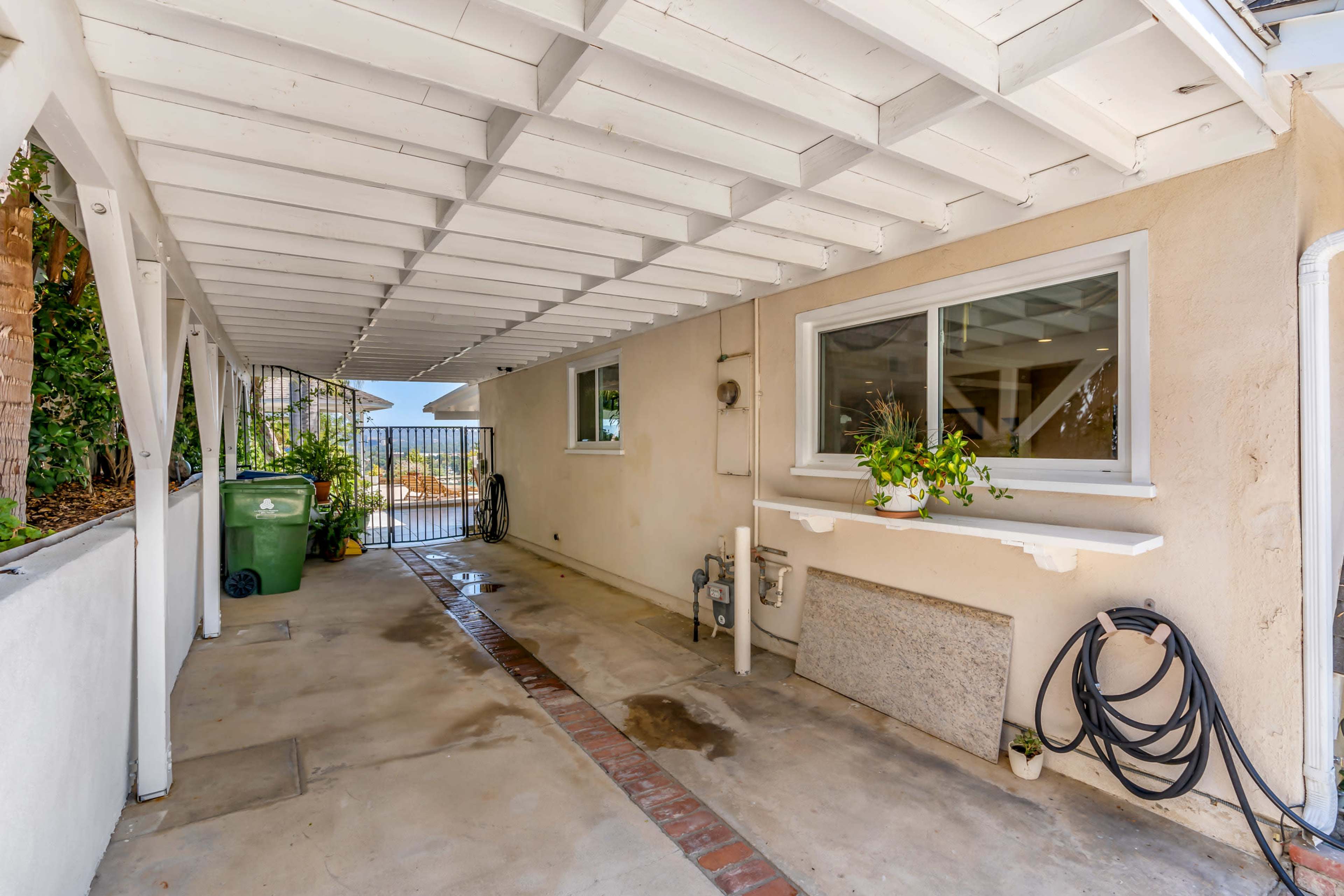 A covered walkway with a concrete floor, a green trash bin, and a gated entrance on one end.